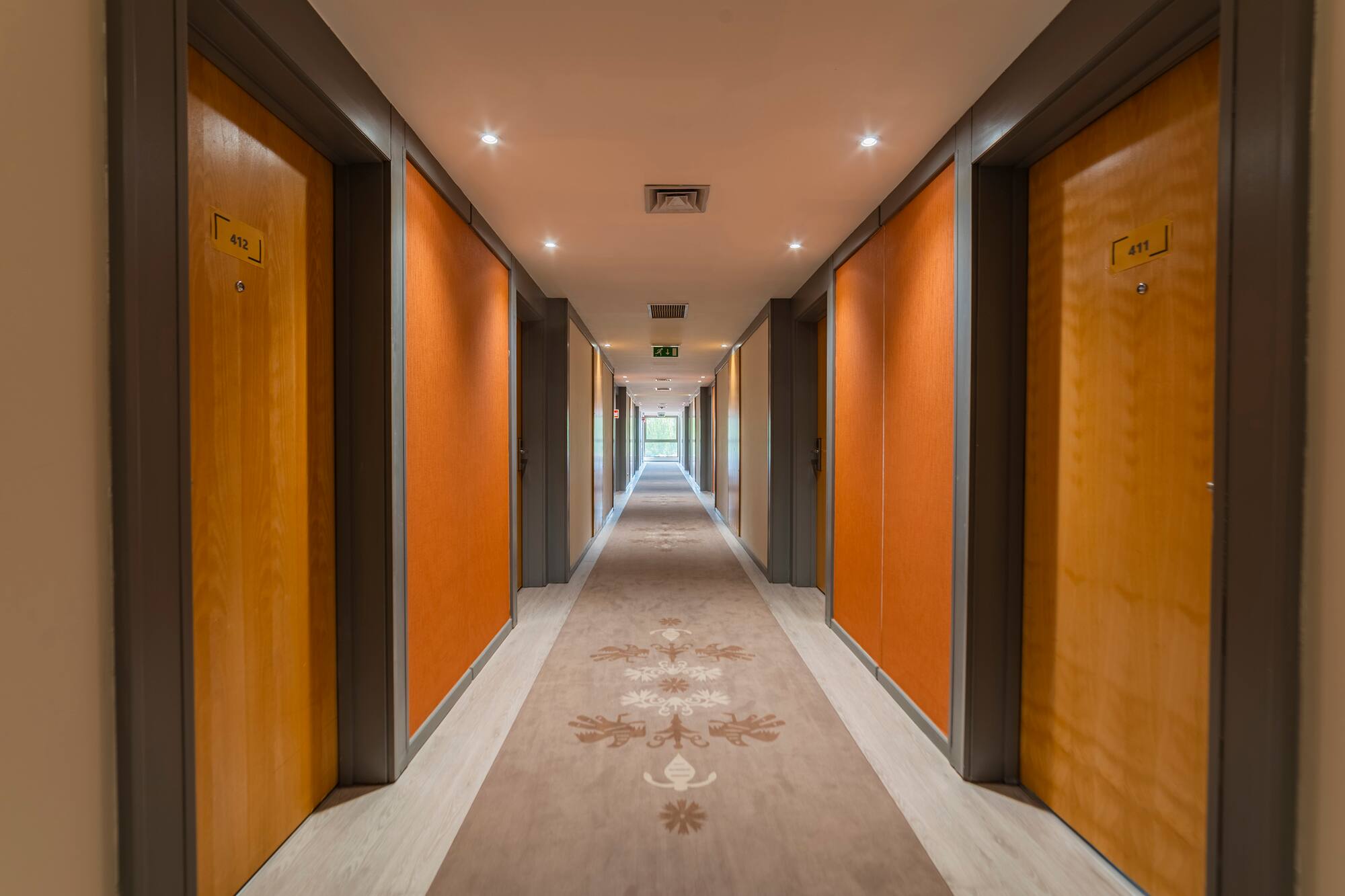Modern hotel corridor with orange walls, patterned carpet, and wooden room doors.