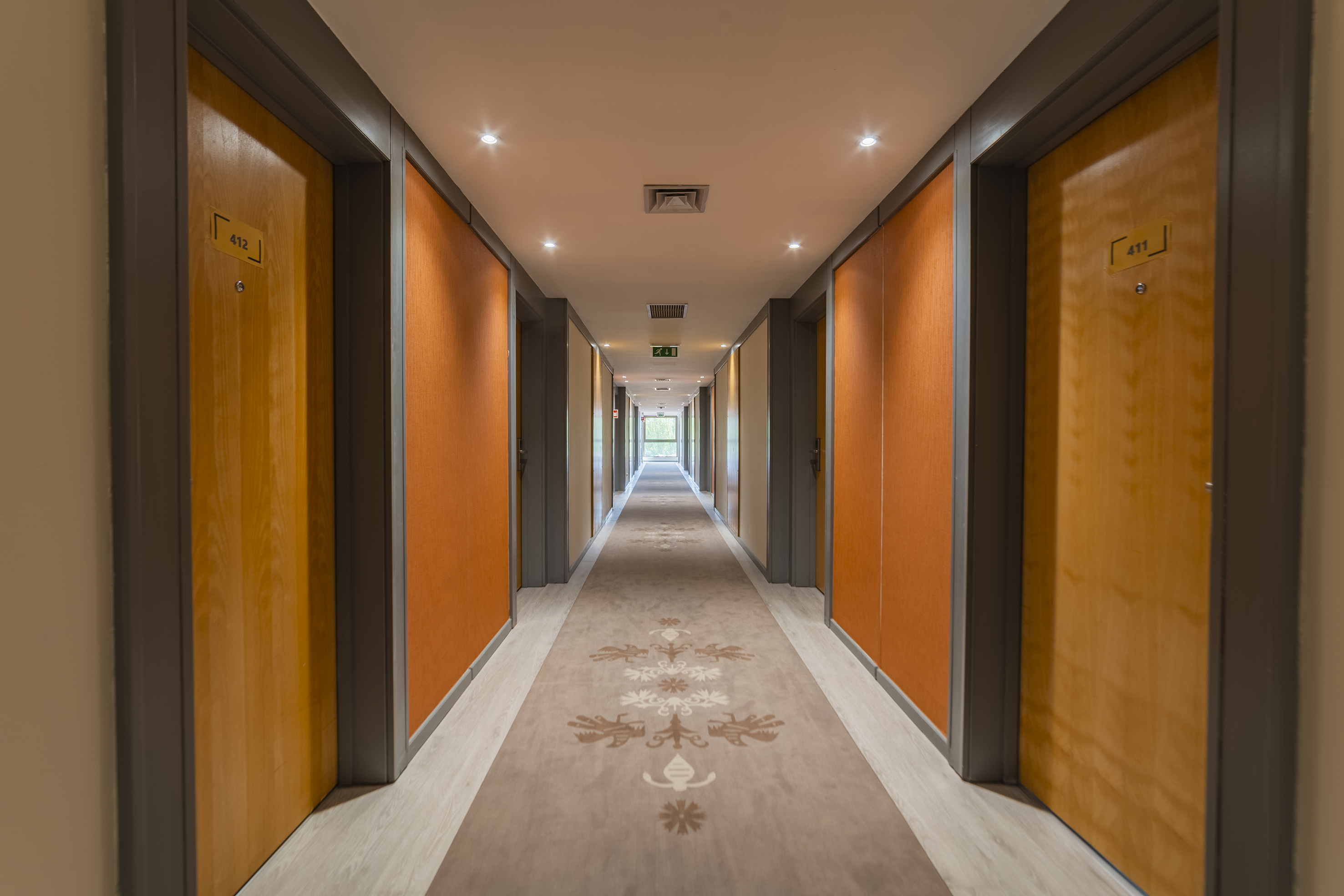 Modern hotel corridor with orange walls, patterned carpet, and wooden room doors.