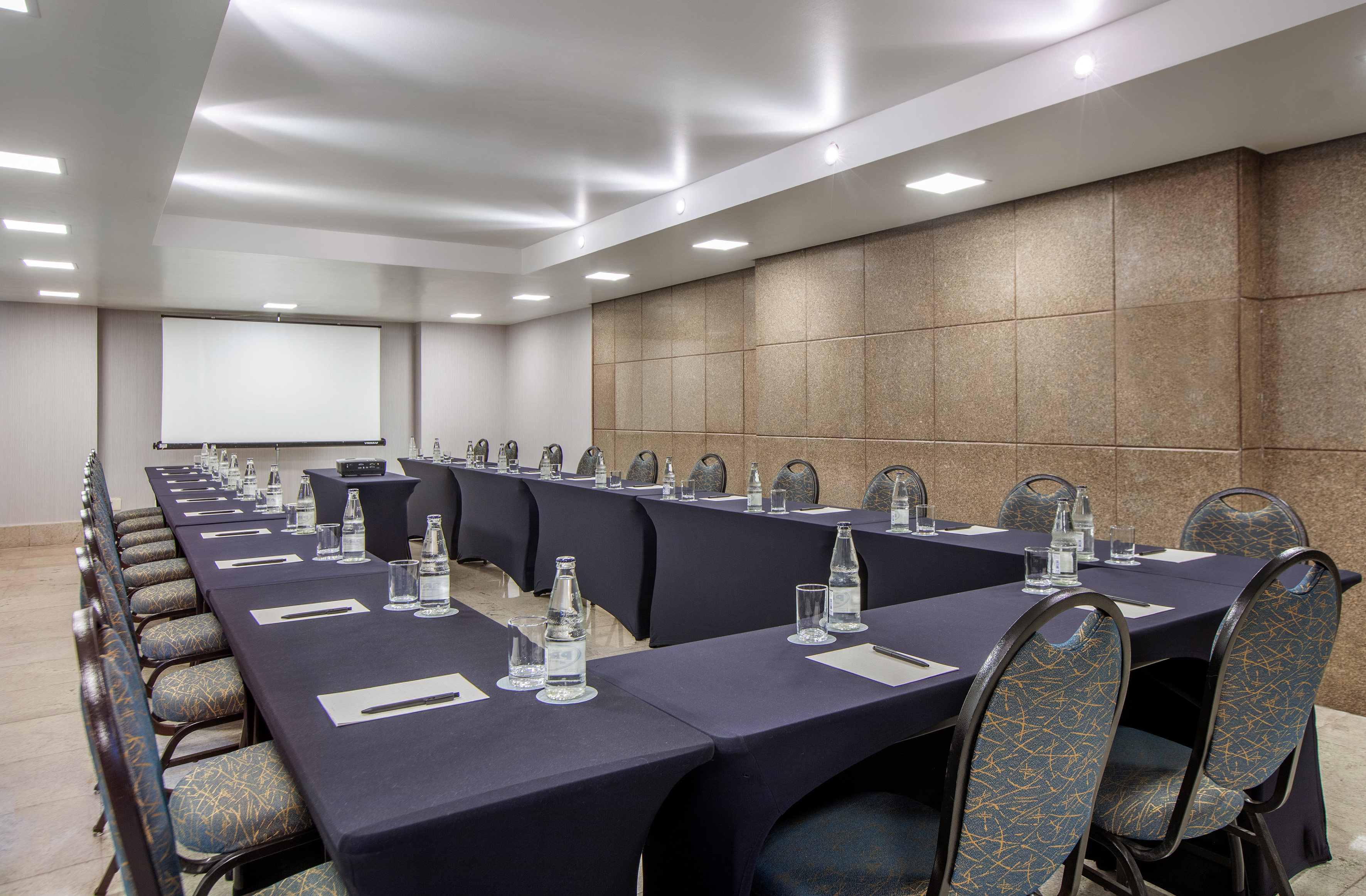 U-shaped meeting room setup with black tables, patterned chairs, and projector screen. - Encontro IV - -Hotel Madrid Airport Suites Affiliated by Meliá:, Brasilia