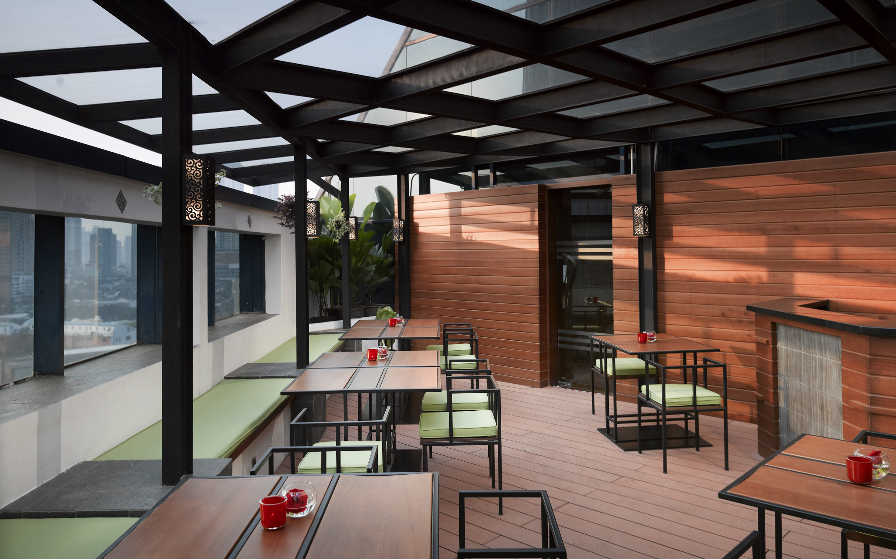 Modern rooftop dining terrace with wooden tables, green cushions, and city view.