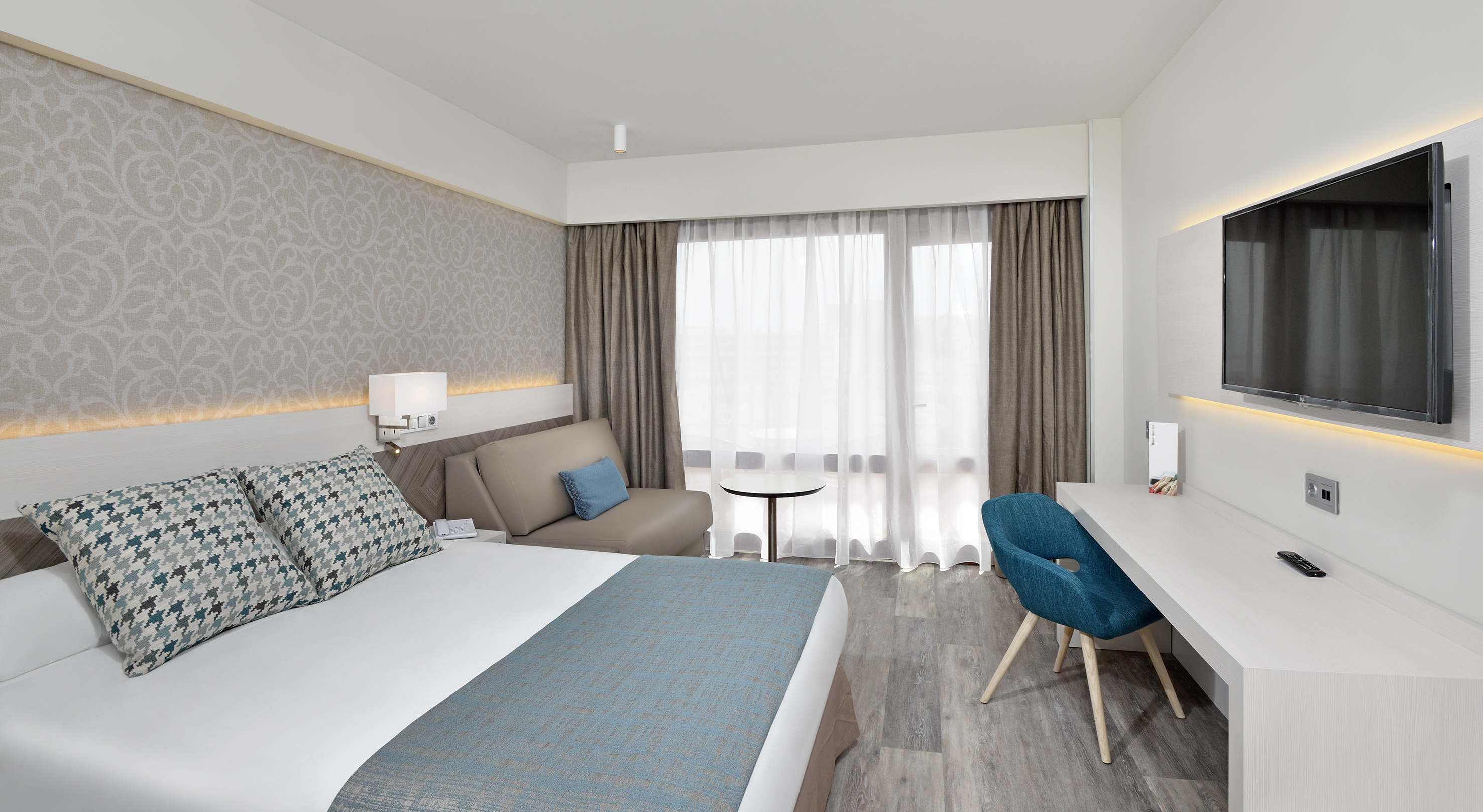 Modern hotel room with bed, blue chair, desk, wall-mounted TV, and bright window.