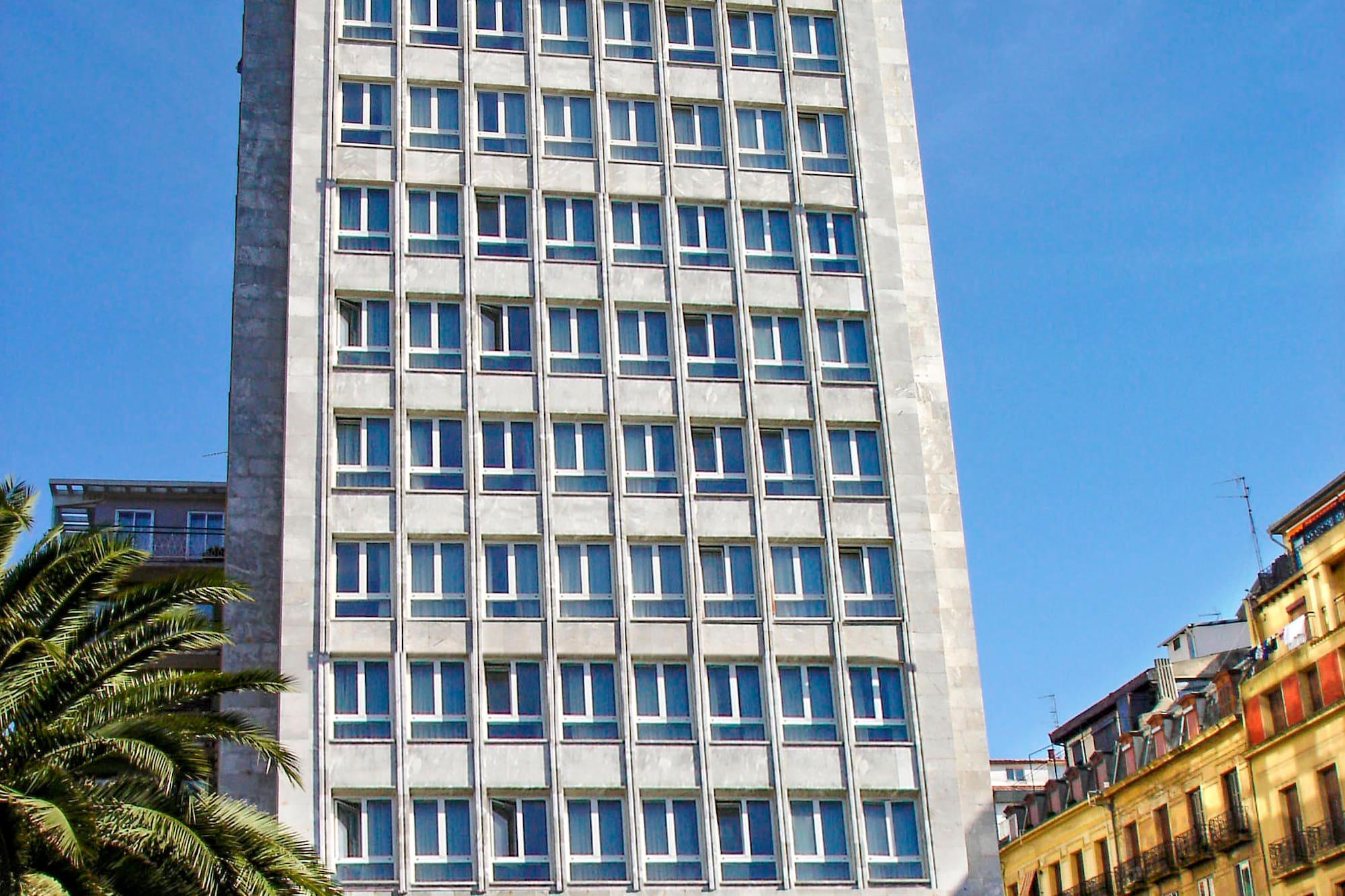 Tall grey modern high-rise building with rows of windows, palm trees, urban street.