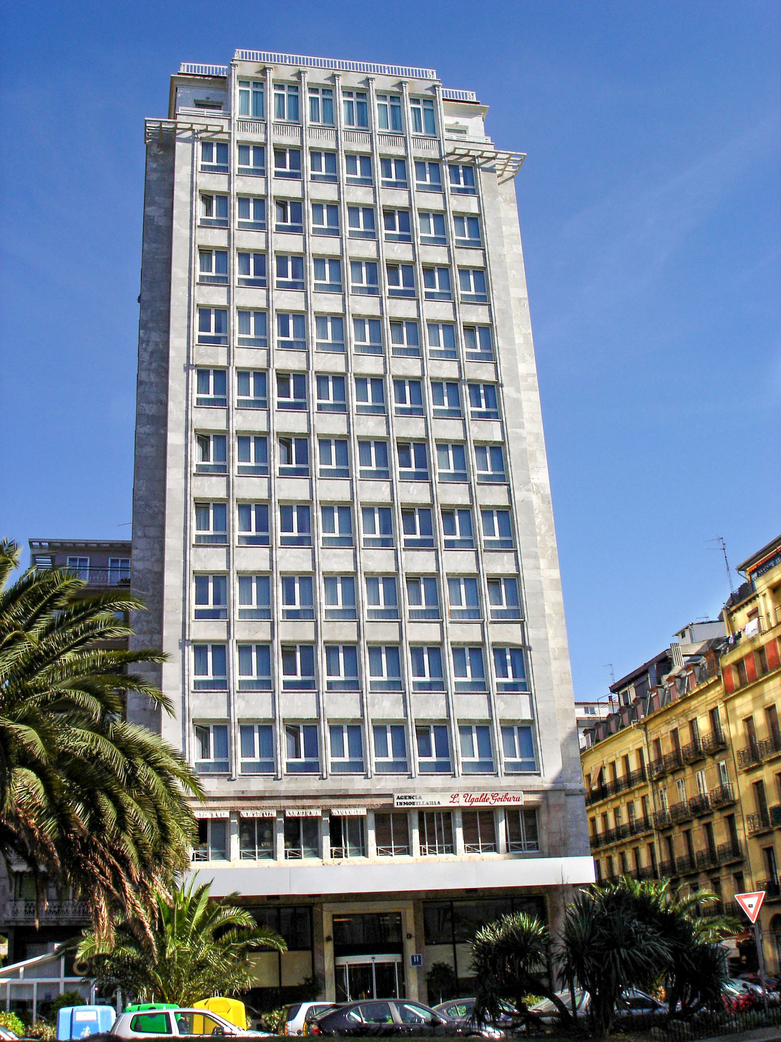 Tall grey modern high-rise building with rows of windows, palm trees, urban street.
