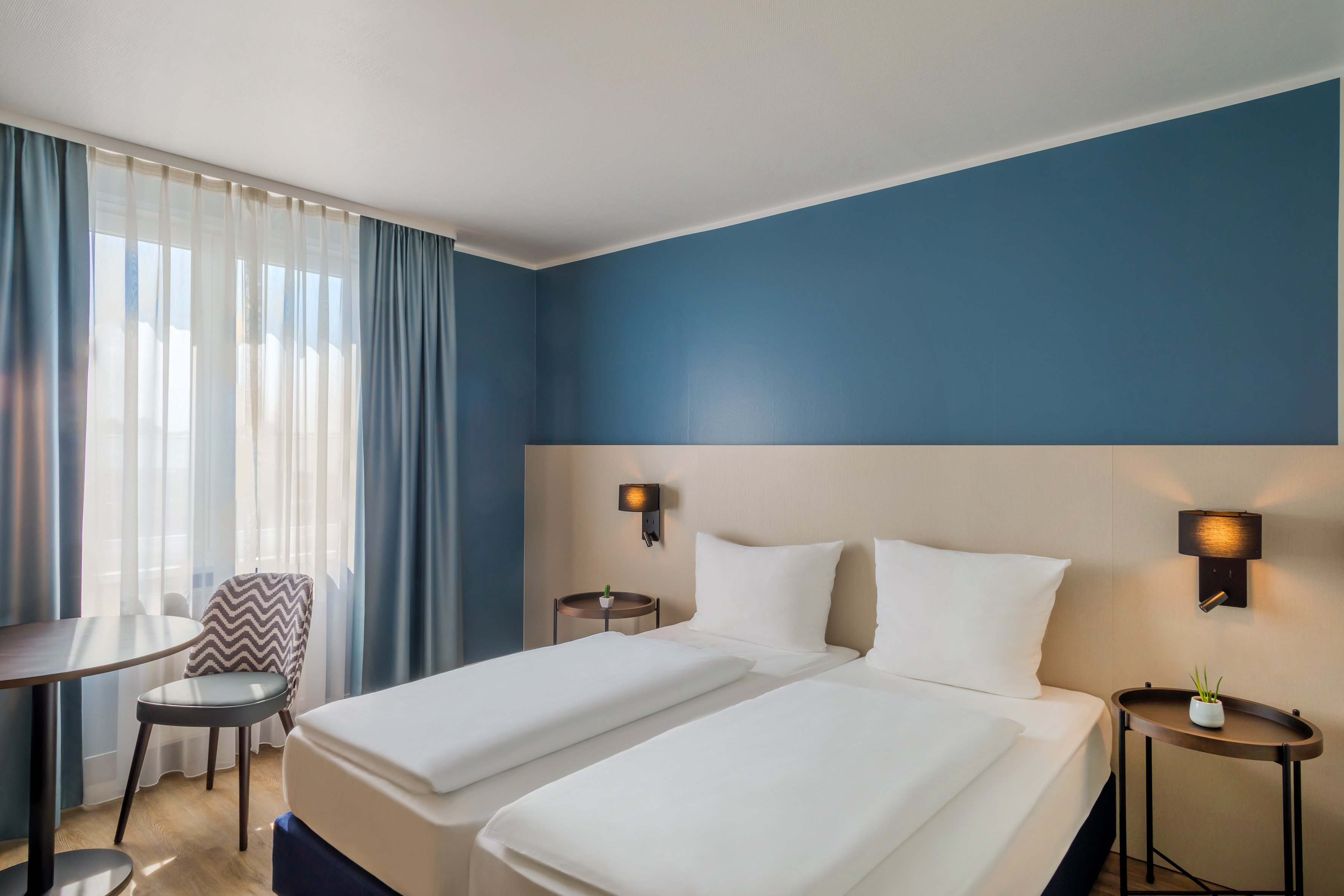 Modern hotel room with two single beds, blue accent wall, and window.