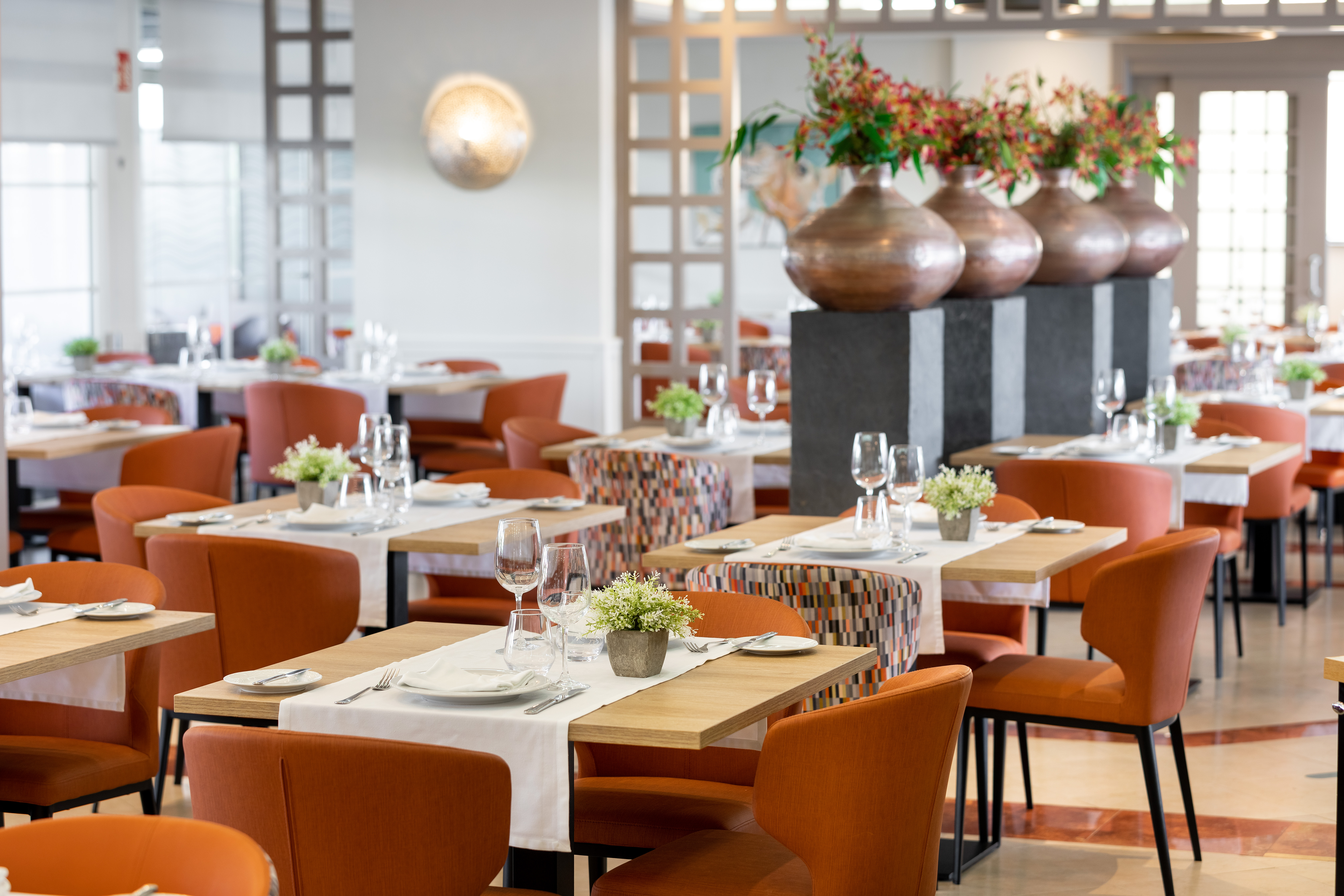 Modern restaurant dining room with set tables, orange chairs, and floral decor.