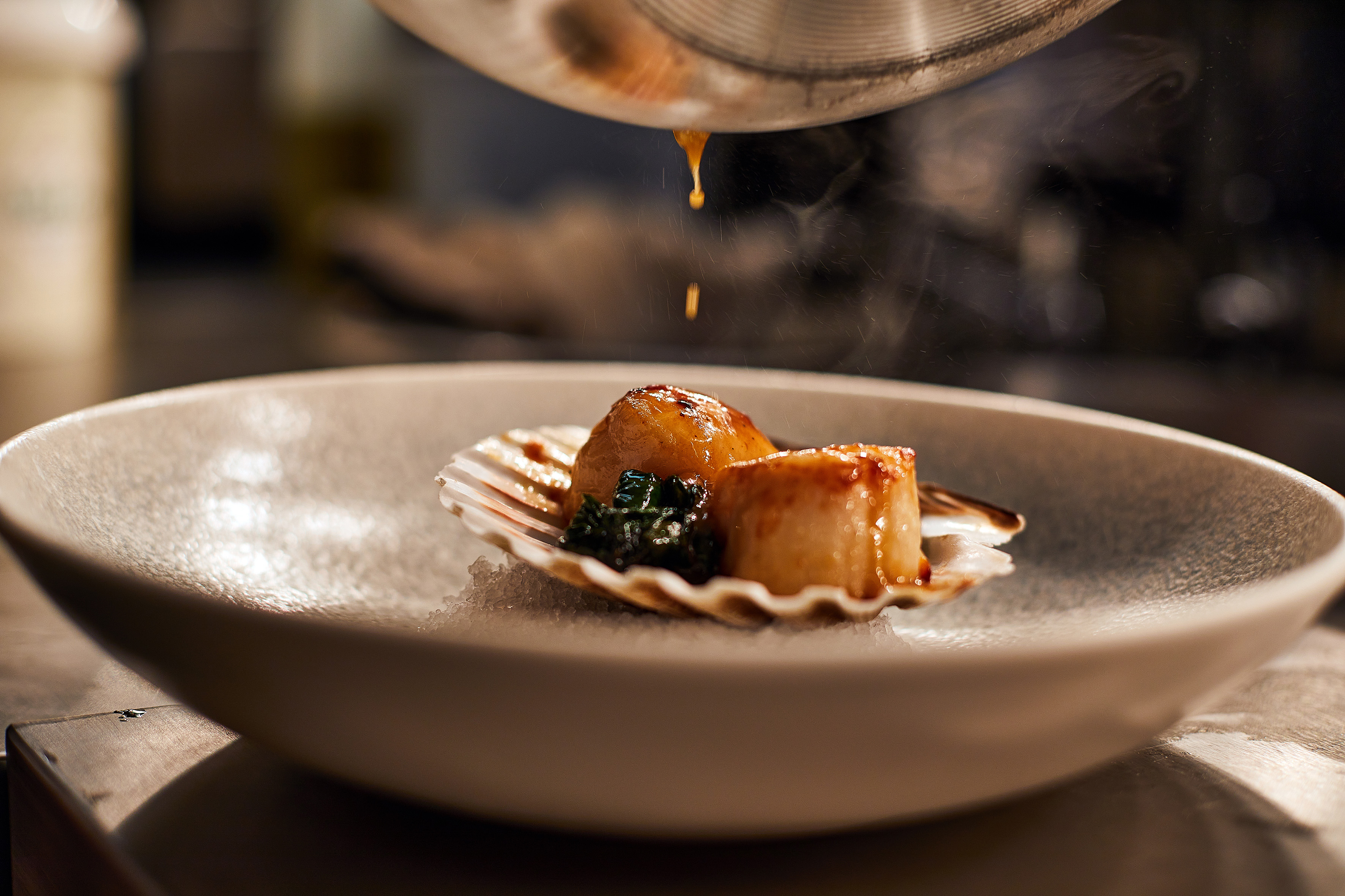 Sauce drizzling over seared scallops in a shell, gourmet seafood presentation.
