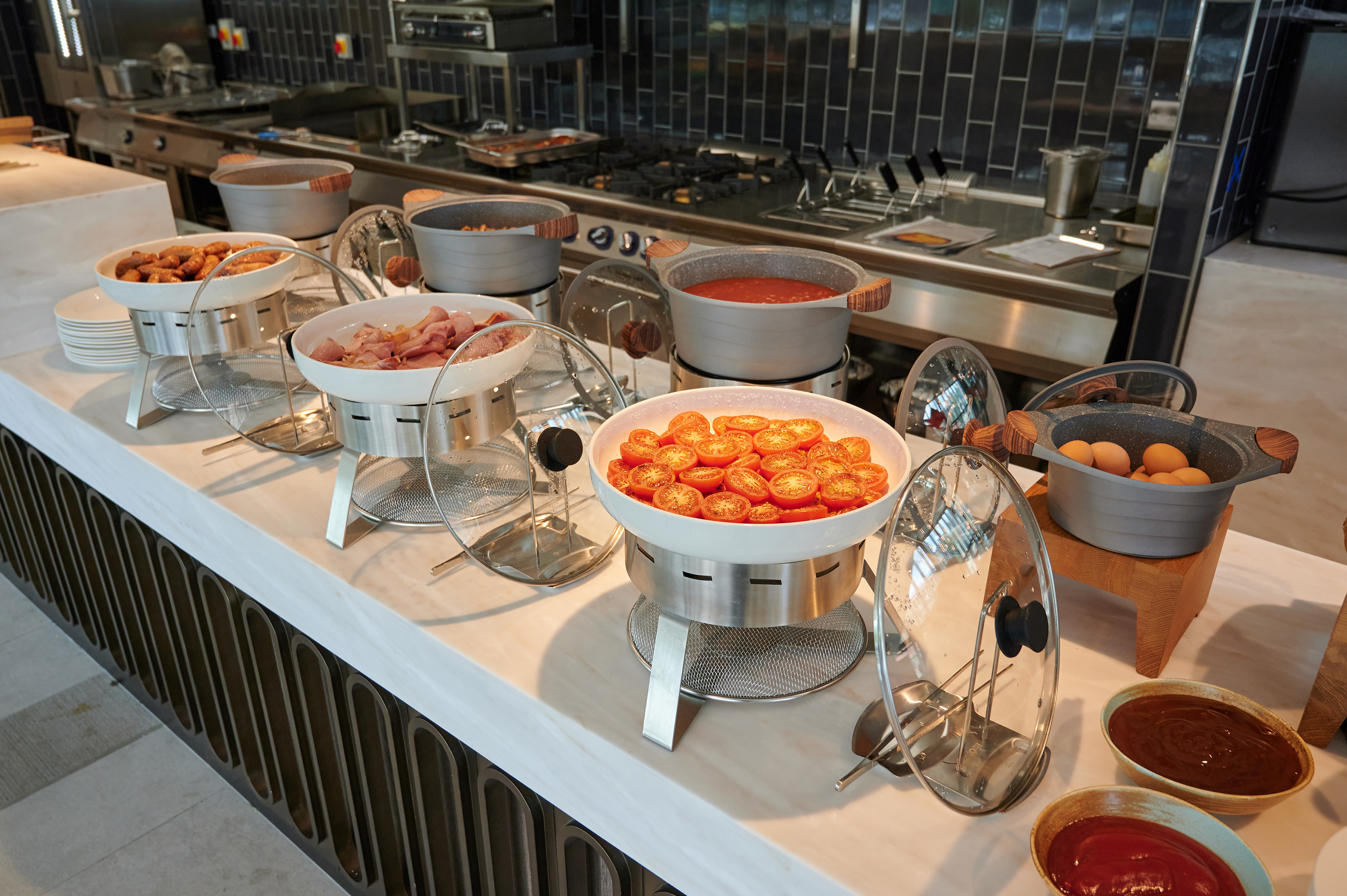 Hotel breakfast buffet with hot sausages, ham, sliced tomatoes, and eggs.