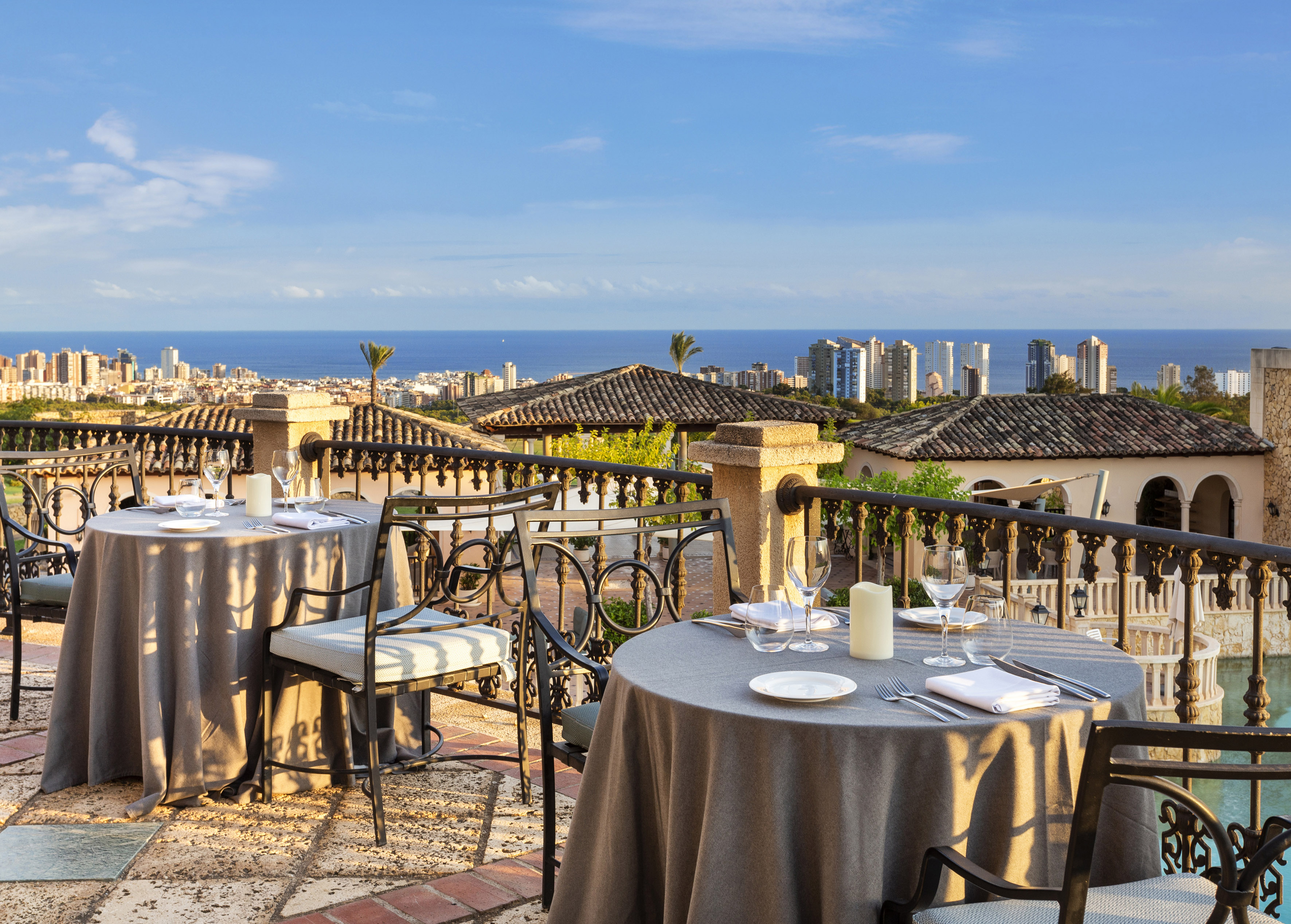 Outdoor restaurant terrace with set tables, ocean view, and coastal city skyline.