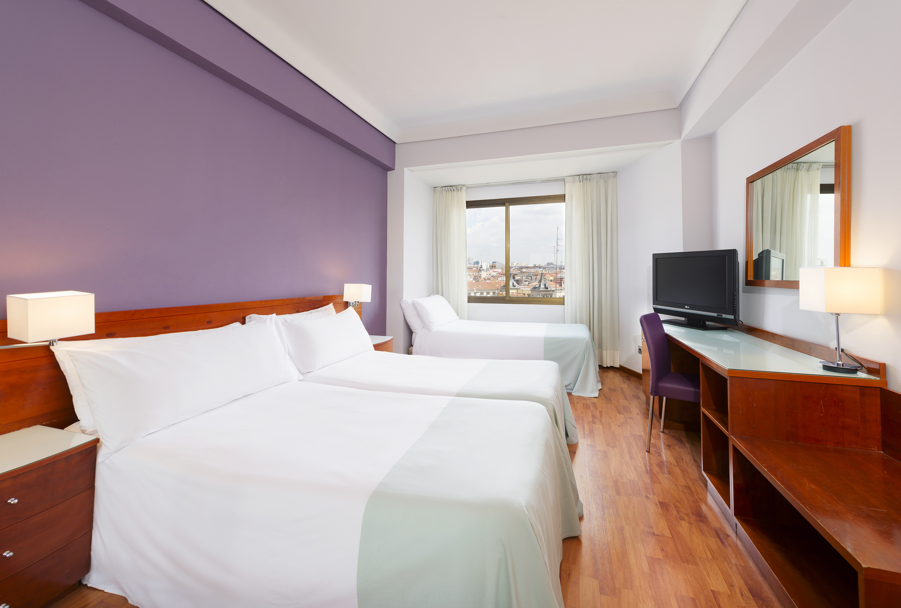 Family hotel room with three beds, purple wall, desk, and city view.