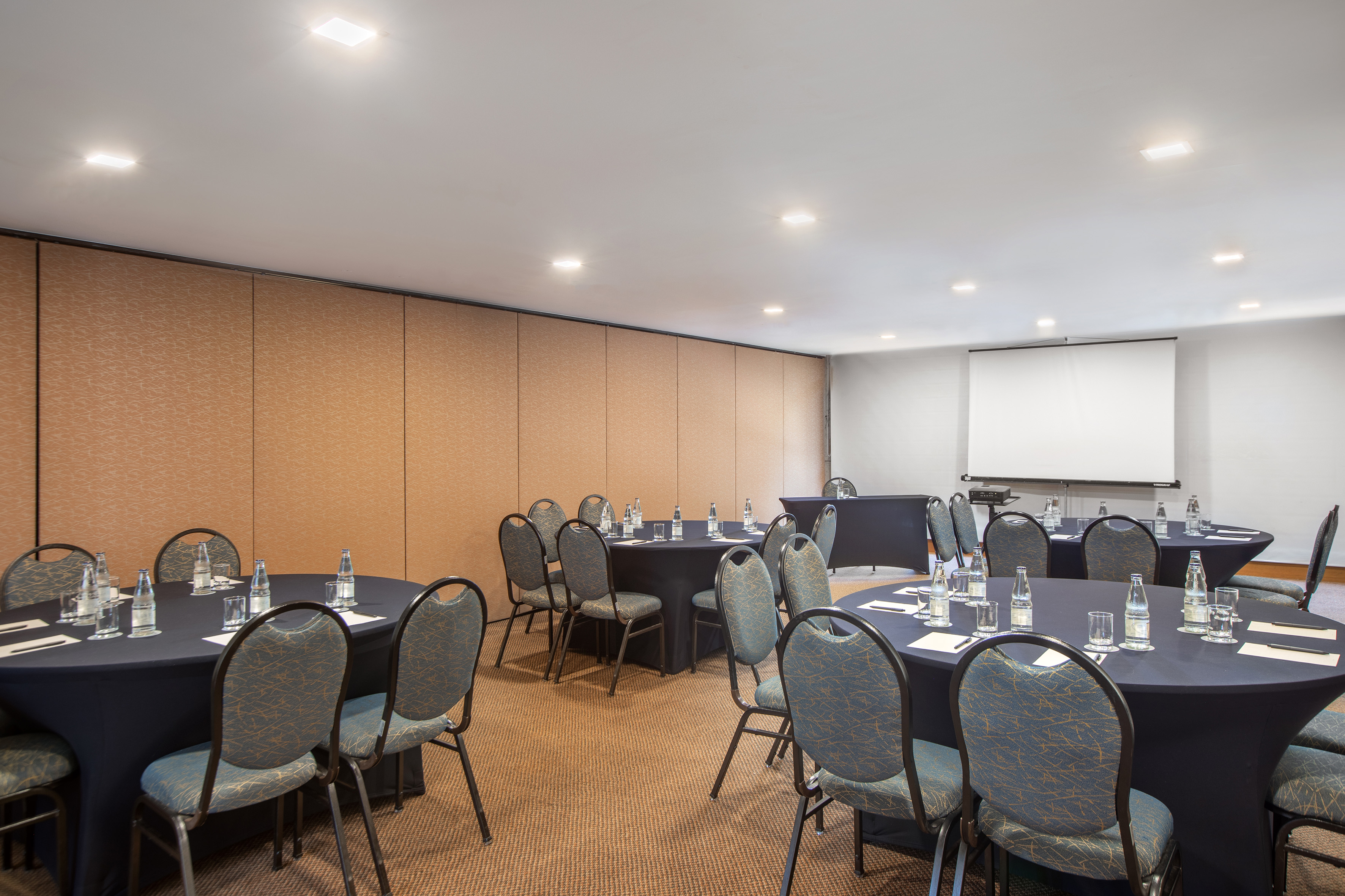 Spacious meeting room with round tables, chairs, water, and projector screen. - Encontro I - -Hotel Madrid Airport Suites Affiliated by Meliá:, Brasilia