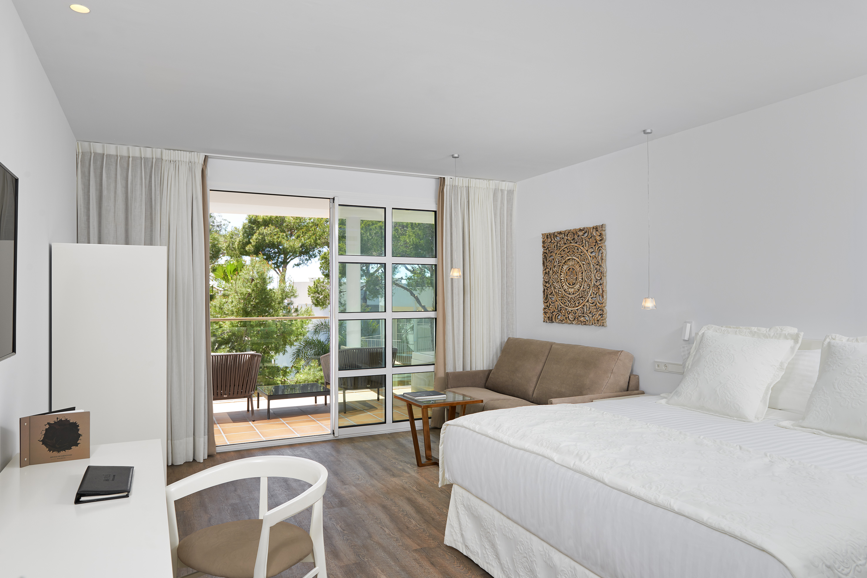 Modern hotel room with king bed, sofa, and balcony overlooking trees.