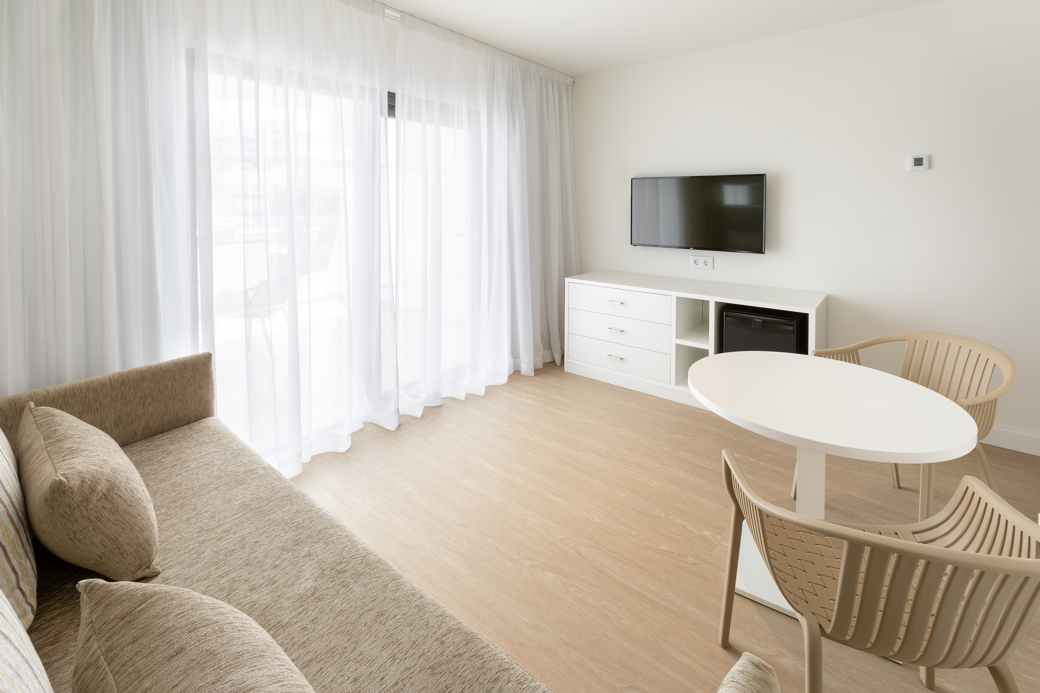 Modern hotel living space with sofa, TV, dining table, and sheer curtains.