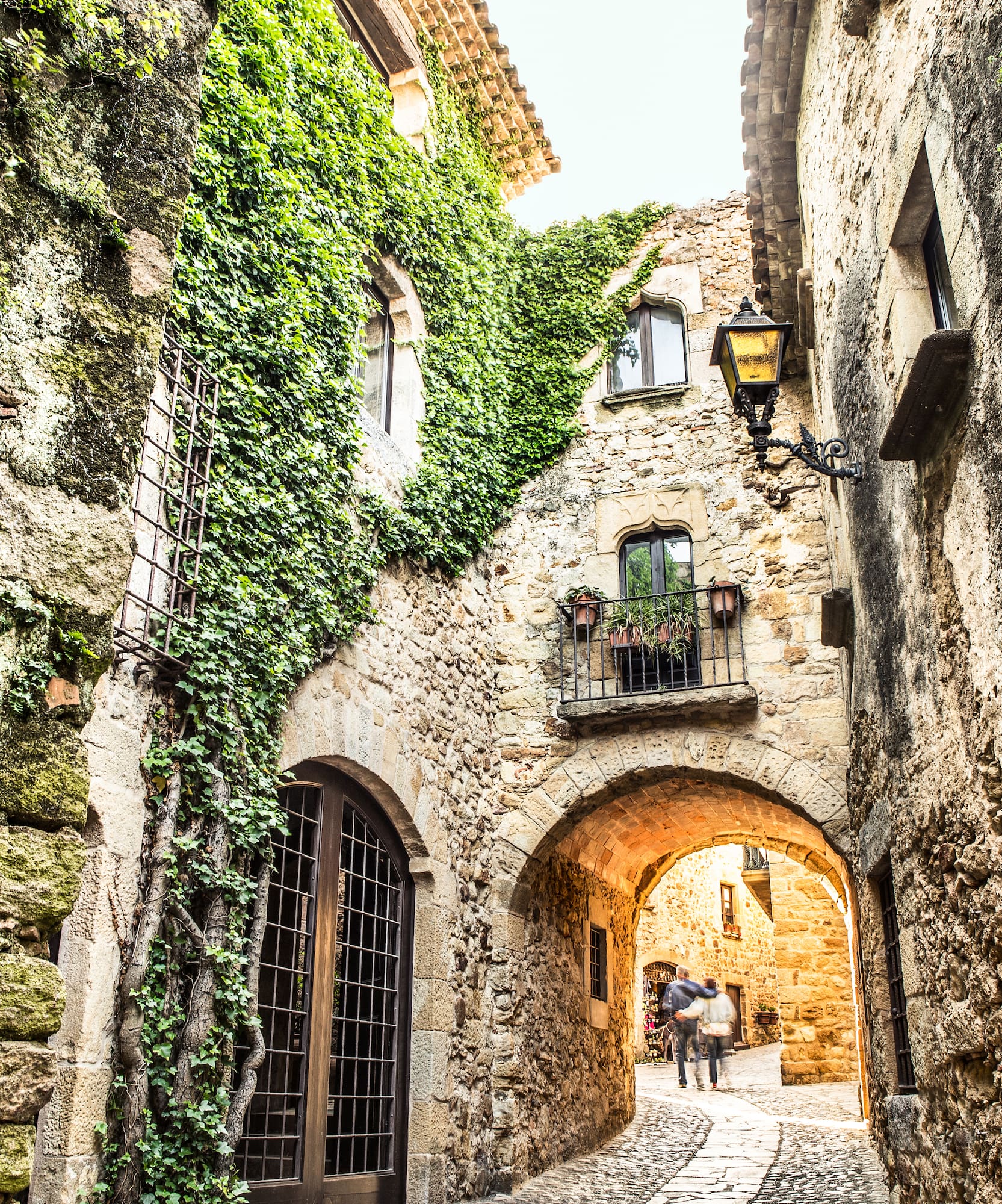 Callejón empedrado medieval con paredes de piedra, hiedra y un arco.