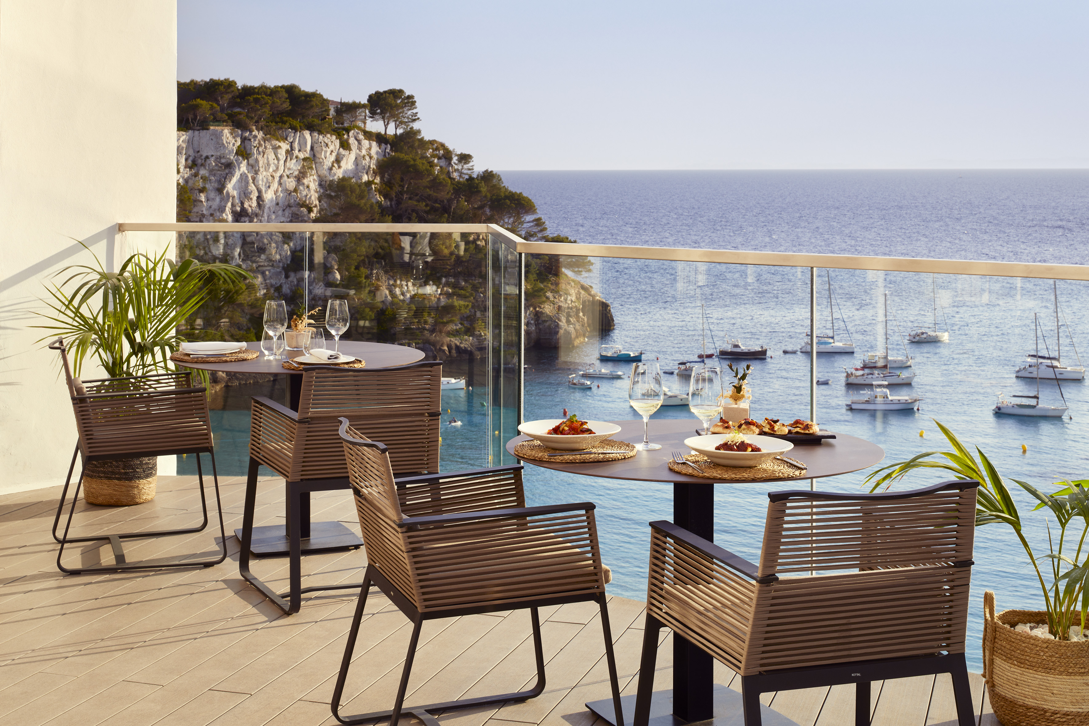 Coastal terrace dining with ocean view, anchored boats, and rocky cliffs.