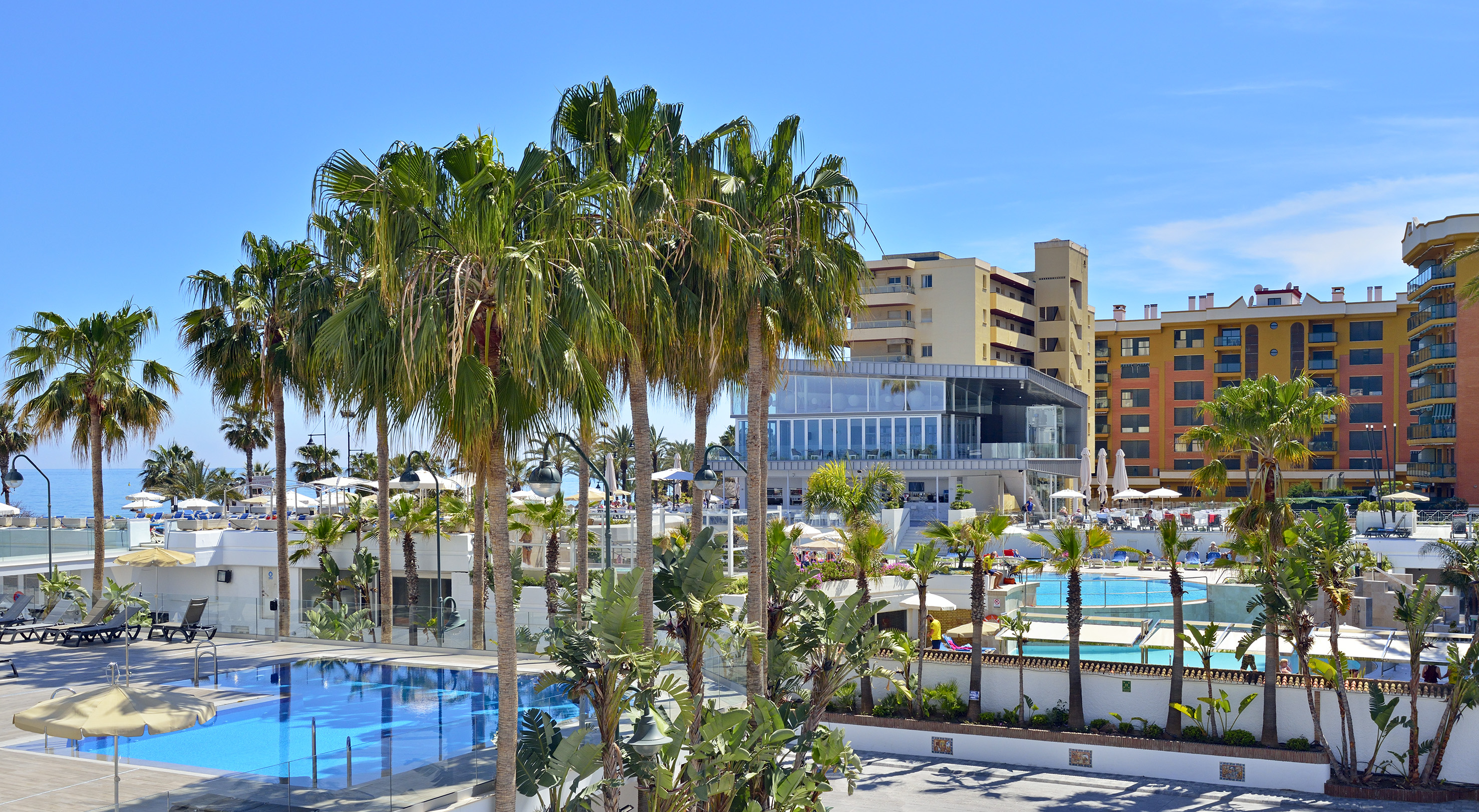 Sunny hotel resort with palm trees, blue swimming pools, and distant ocean.