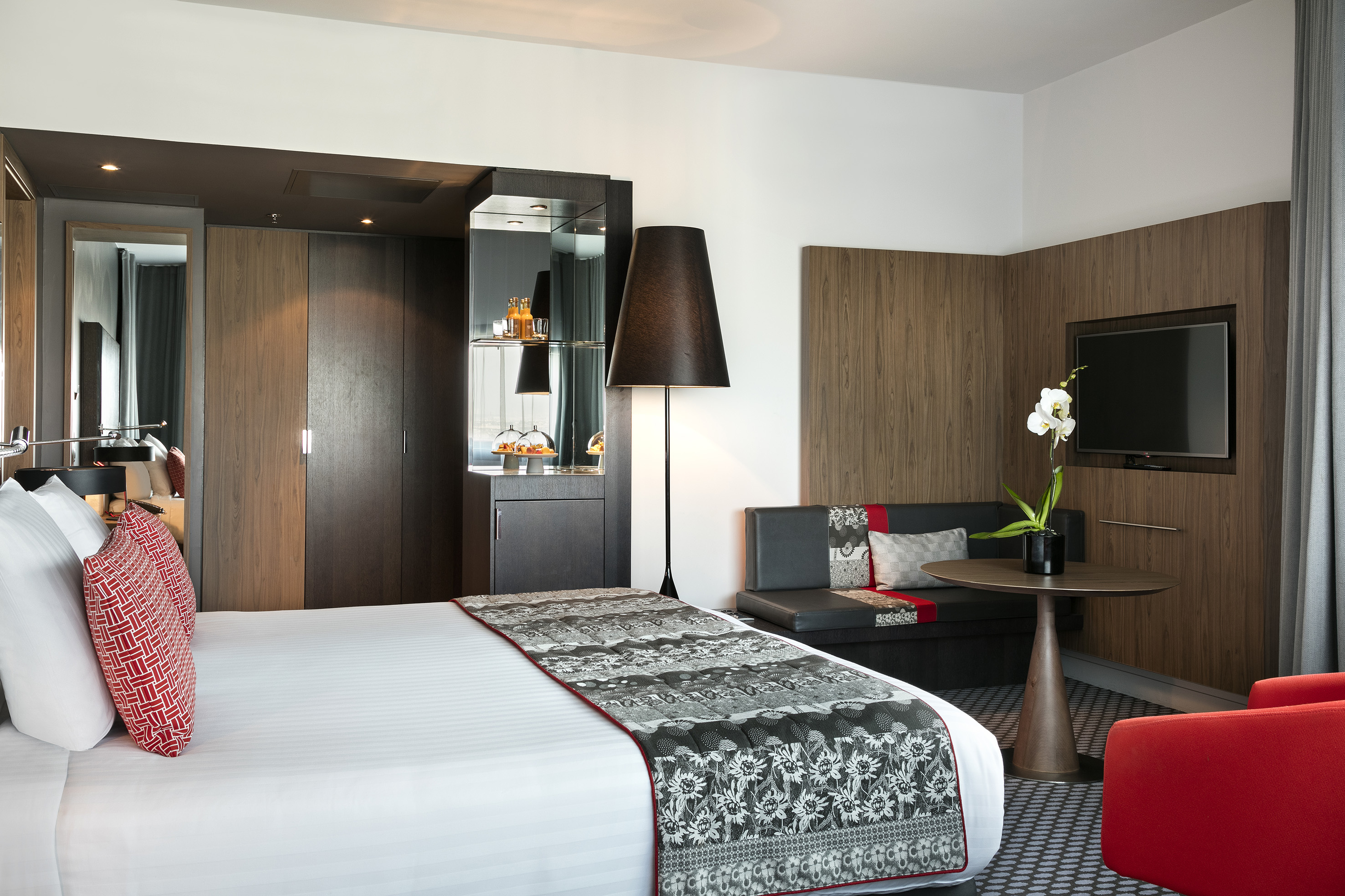 Modern hotel room with large bed, TV, sofa, minibar, and red armchair.