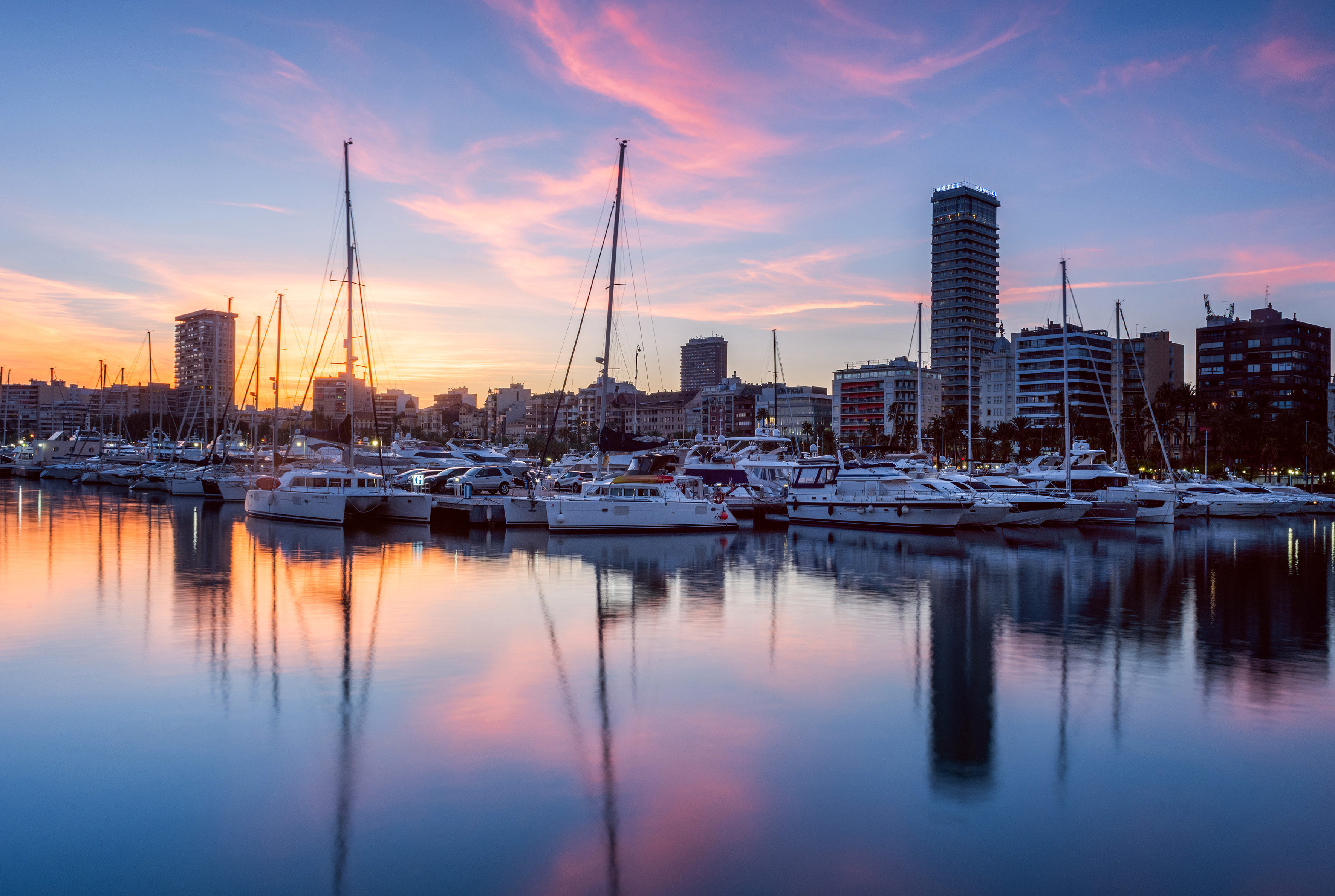 Marina with yachts and city skyline at sunset, reflected in water.