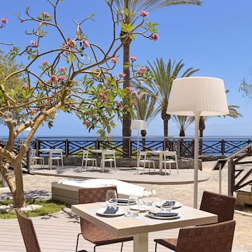 a table and chairs outside with a tree and a beach in the background