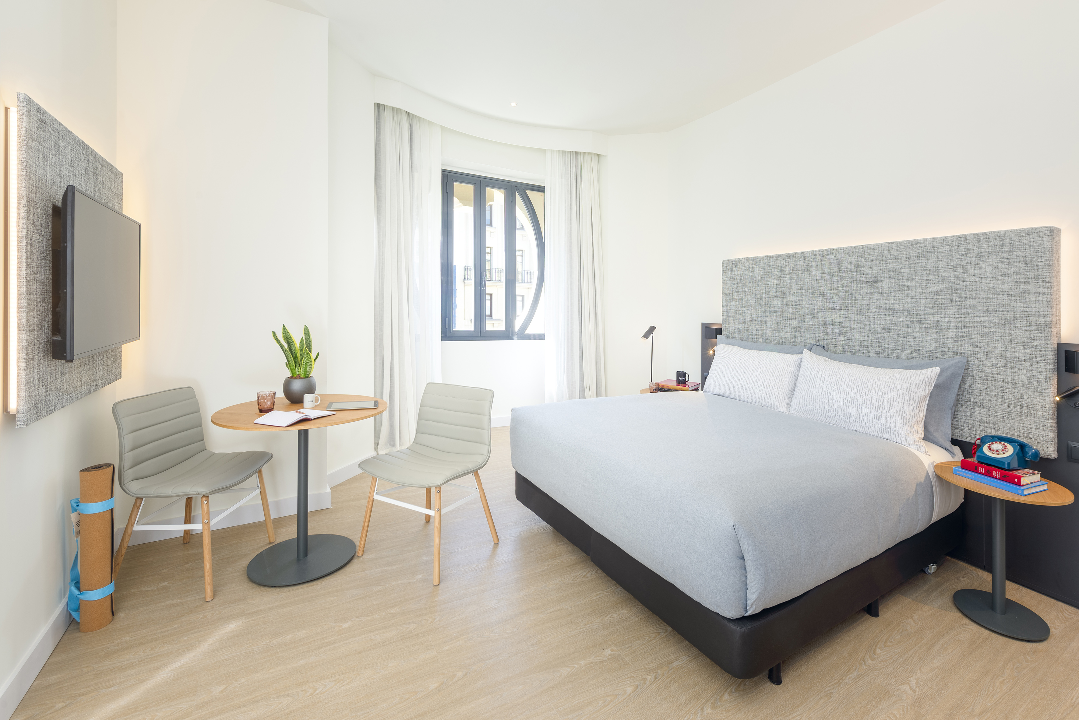Modern hotel bedroom with grey bed, textured headboard, workspace, and TV.