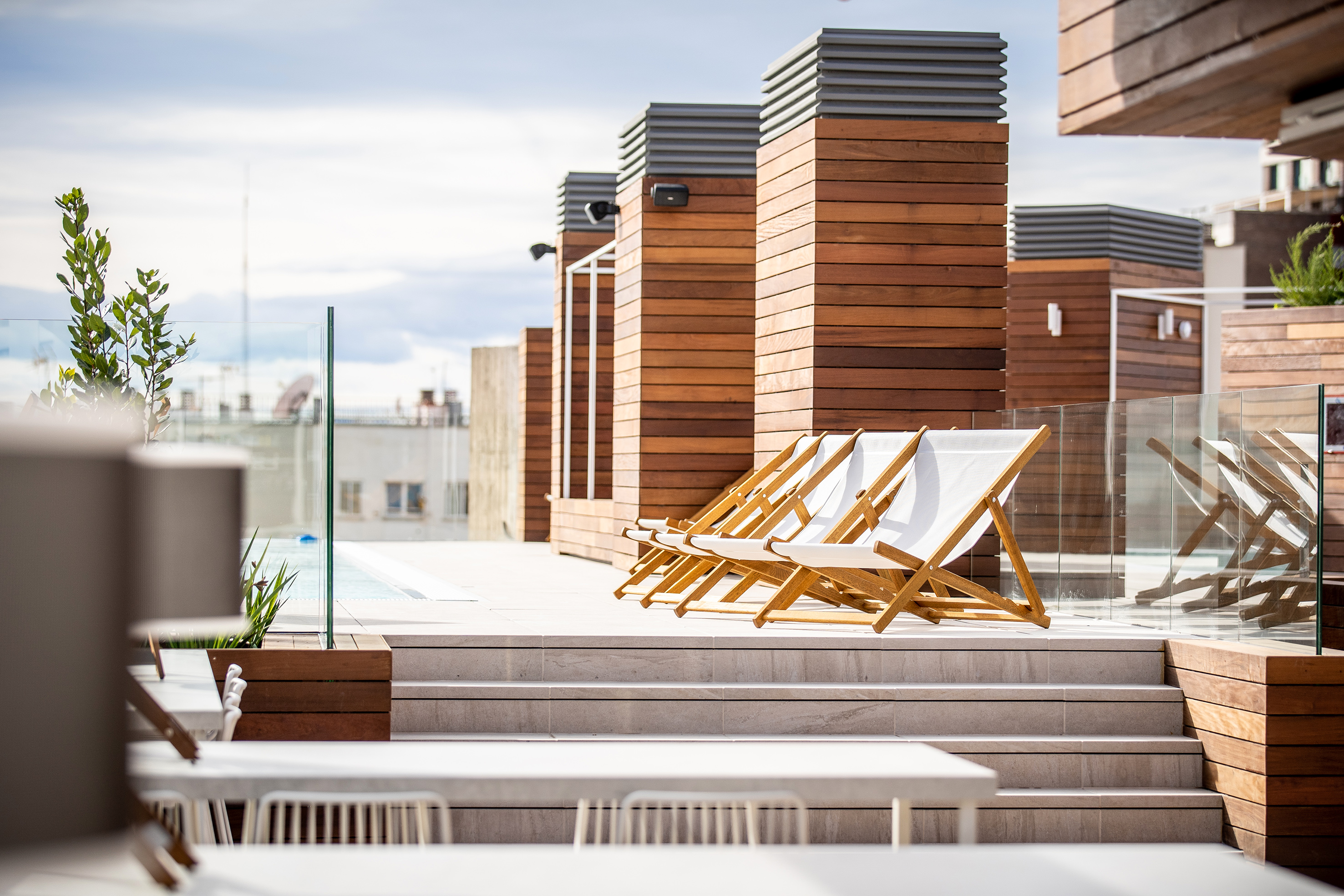 Rooftop pool deck with white lounge chairs, glass railings, and wooden details.