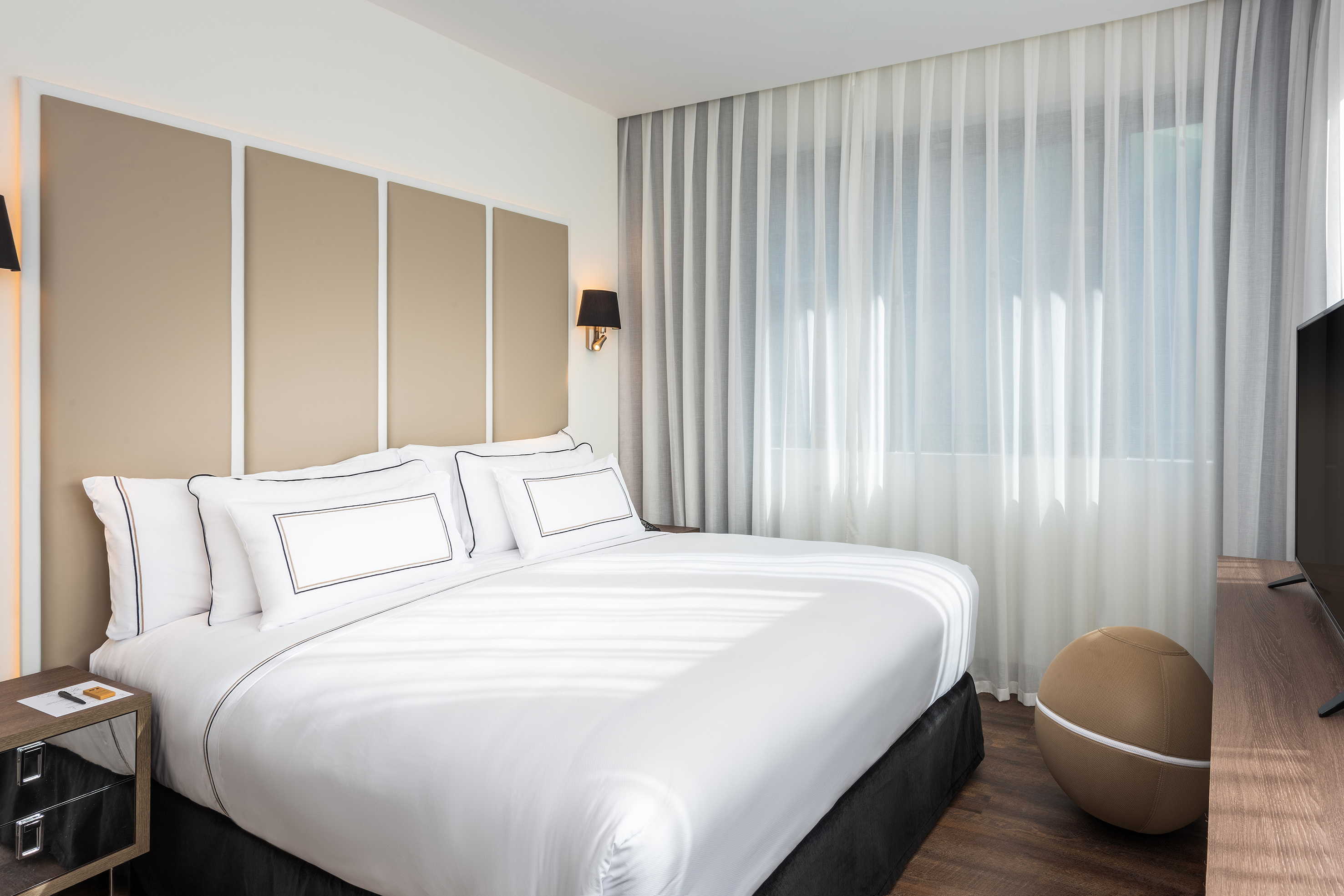 Clean modern hotel bedroom with white king bed, padded headboard, sheer curtains.