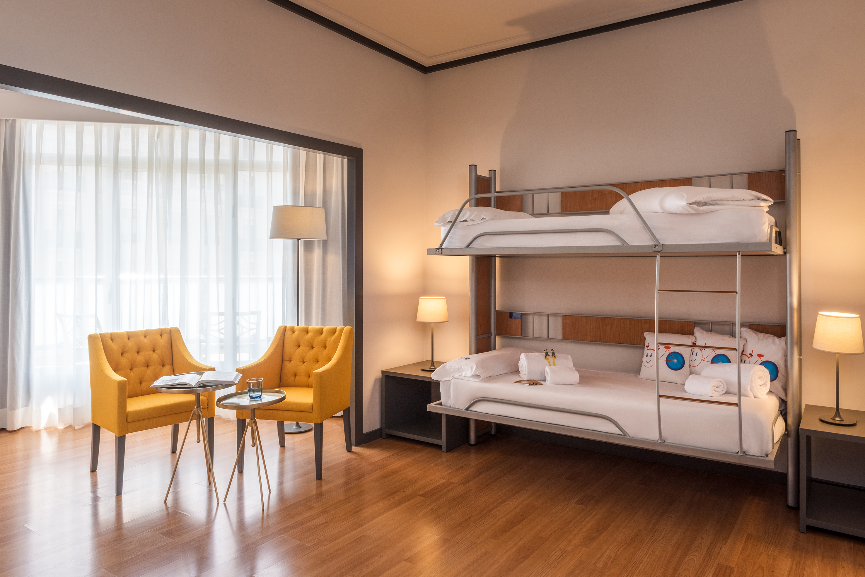 Hotel family room with bunk beds, yellow seating area, and large window.