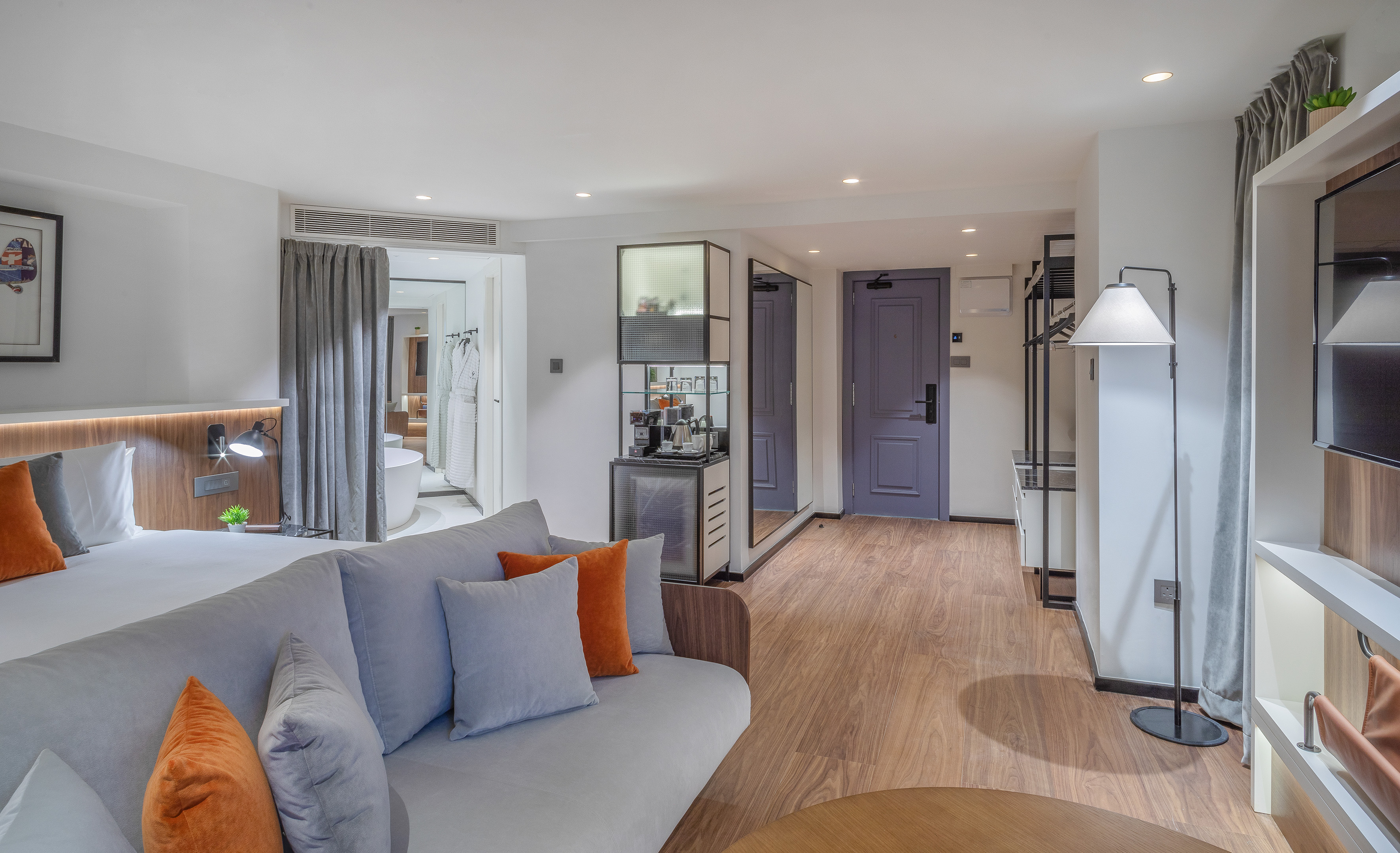 Modern hotel room with bed, gray sofa, wooden floor, bathroom, and minibar.