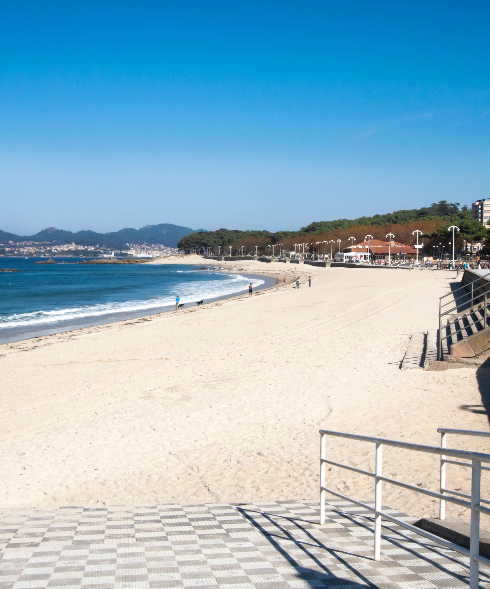 Playa de arena con mar azul, paseo marítimo y ciudad en la distancia, día soleado.