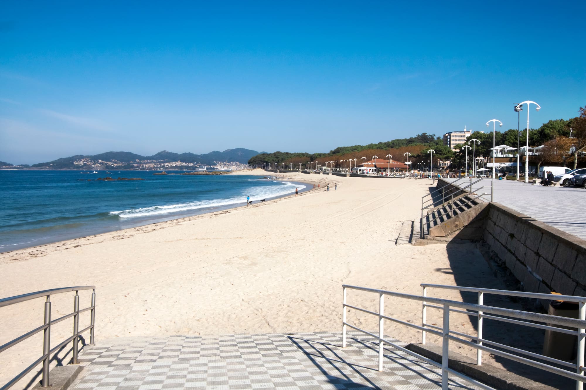 Playa de arena con mar azul, paseo marítimo y ciudad en la distancia, día soleado.