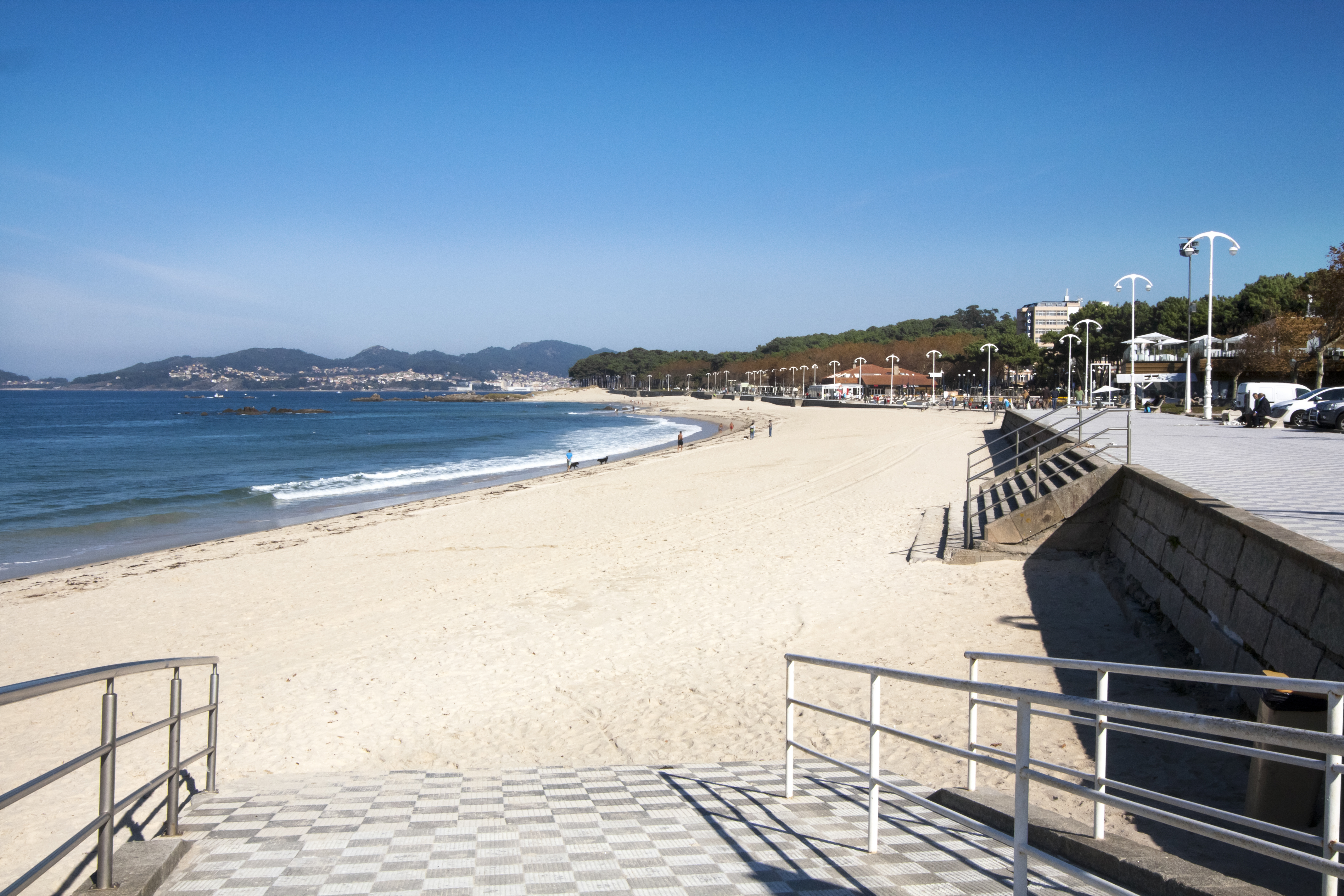 Playa de arena con mar azul, paseo marítimo y ciudad en la distancia, día soleado.