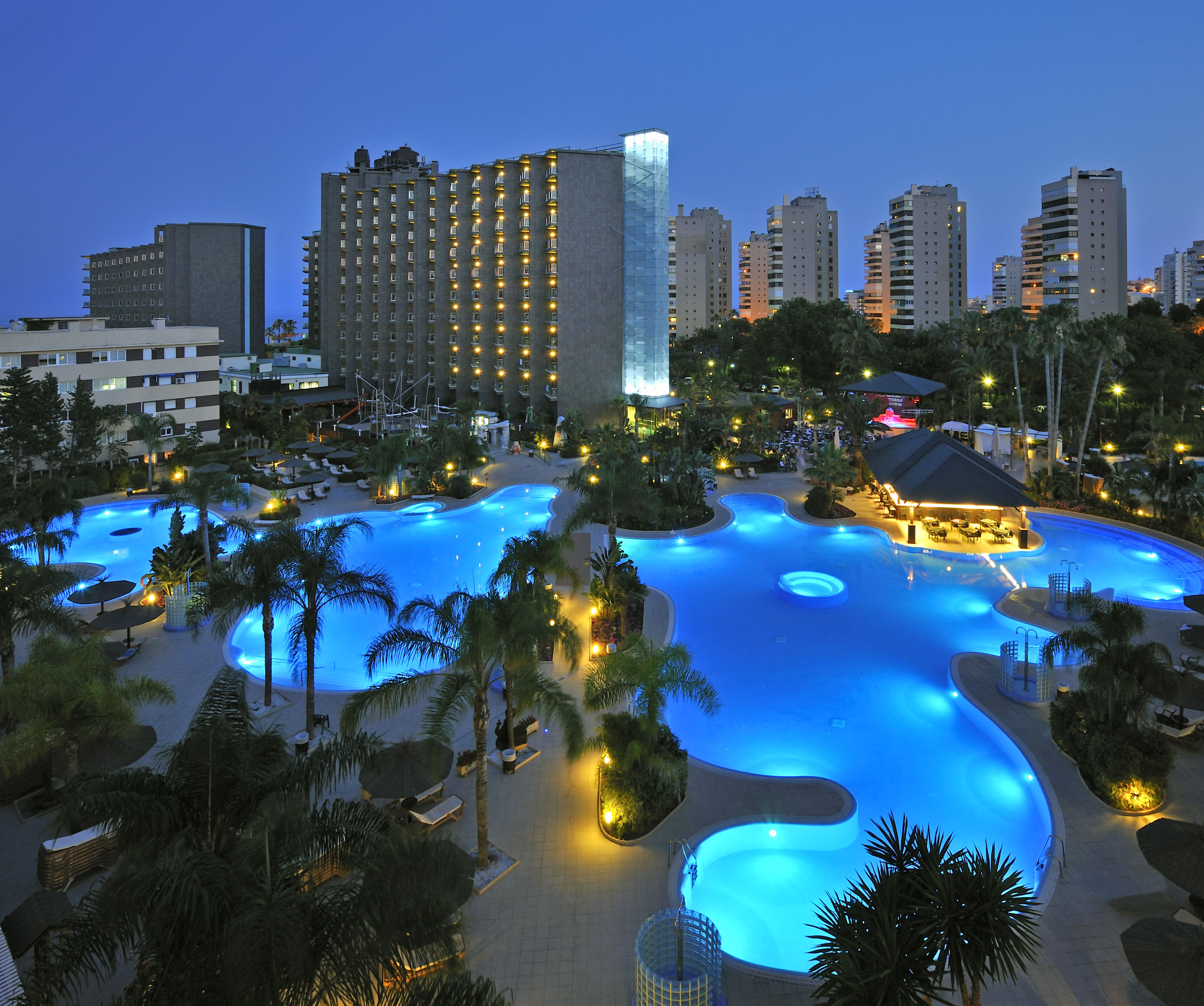 Illuminated resort swimming pool at night with palm trees and hotel buildings.