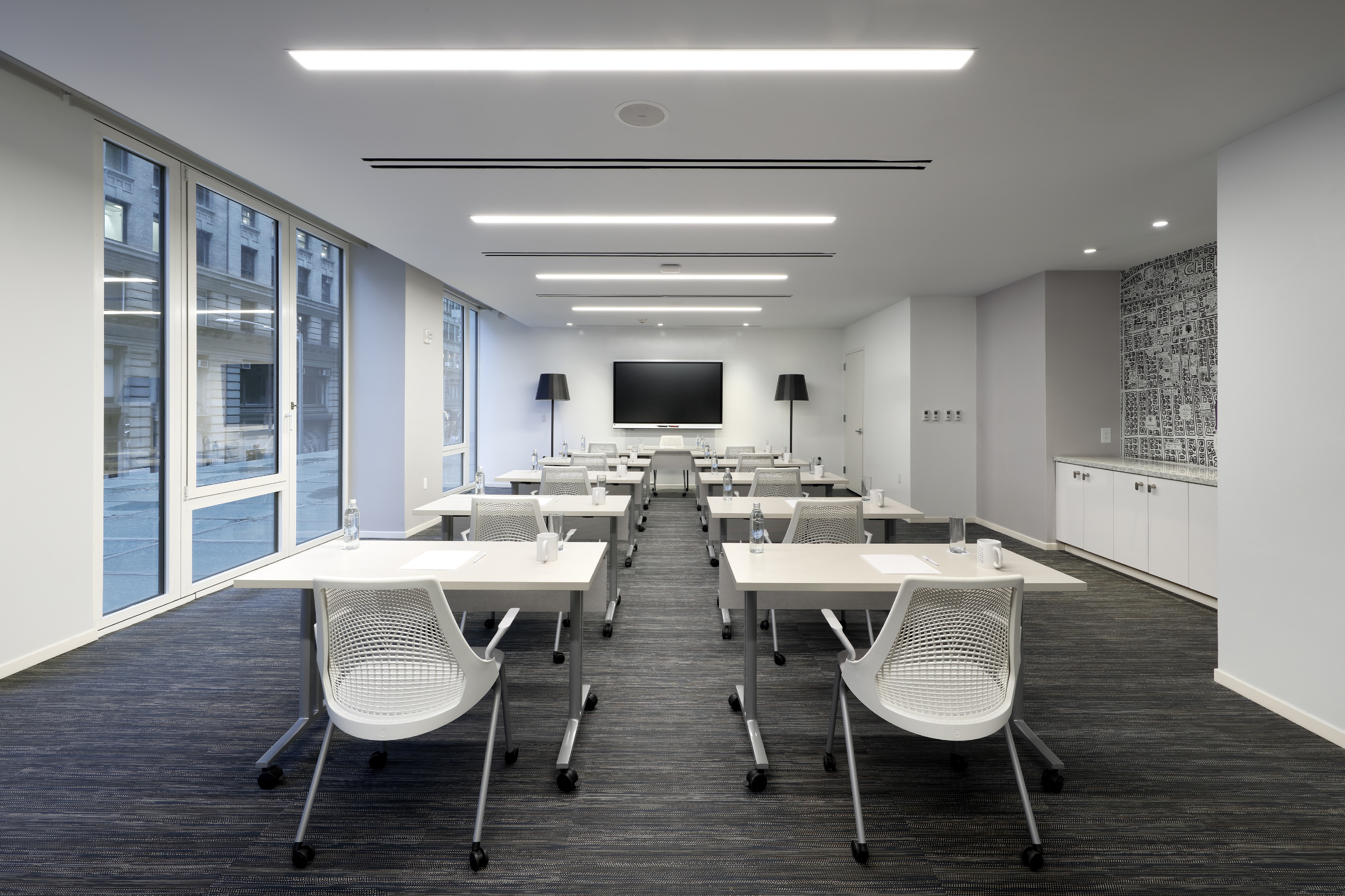 Modern training room with light tables, chairs, large screen, city windows.