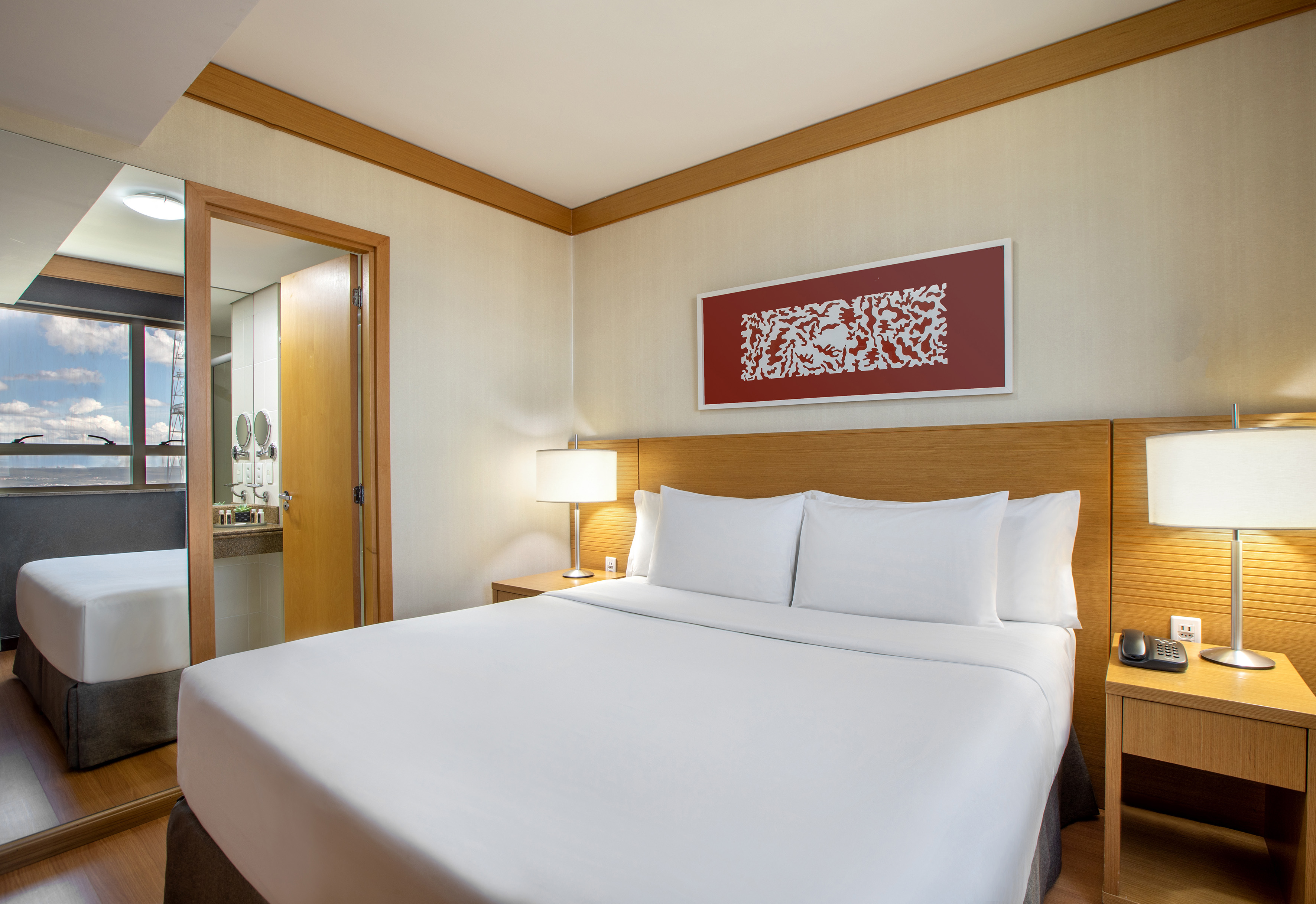 Modern hotel bedroom with king bed, wood furniture, and reflected window view.