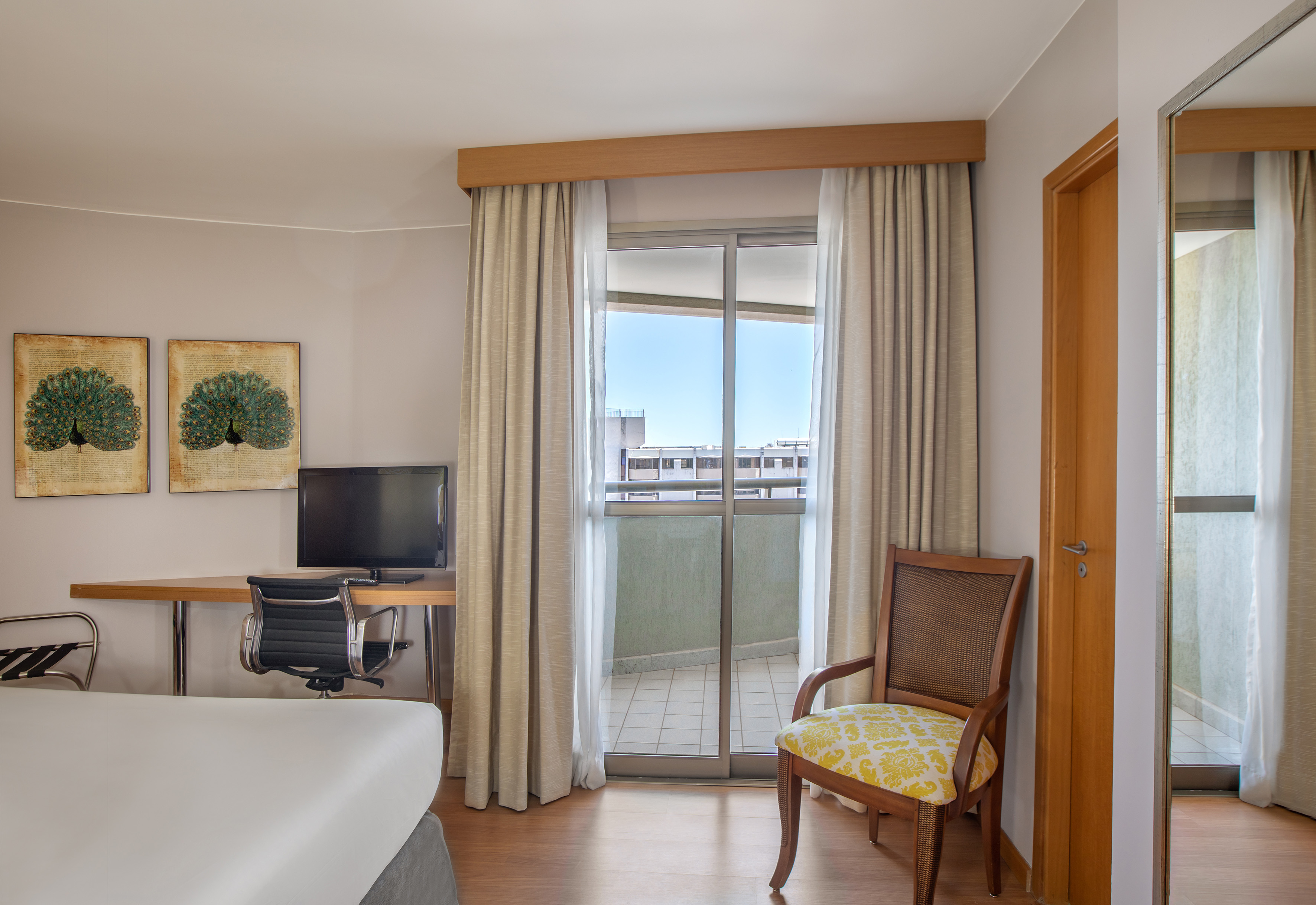 Modern hotel room with balcony, desk, TV, and peacock art.