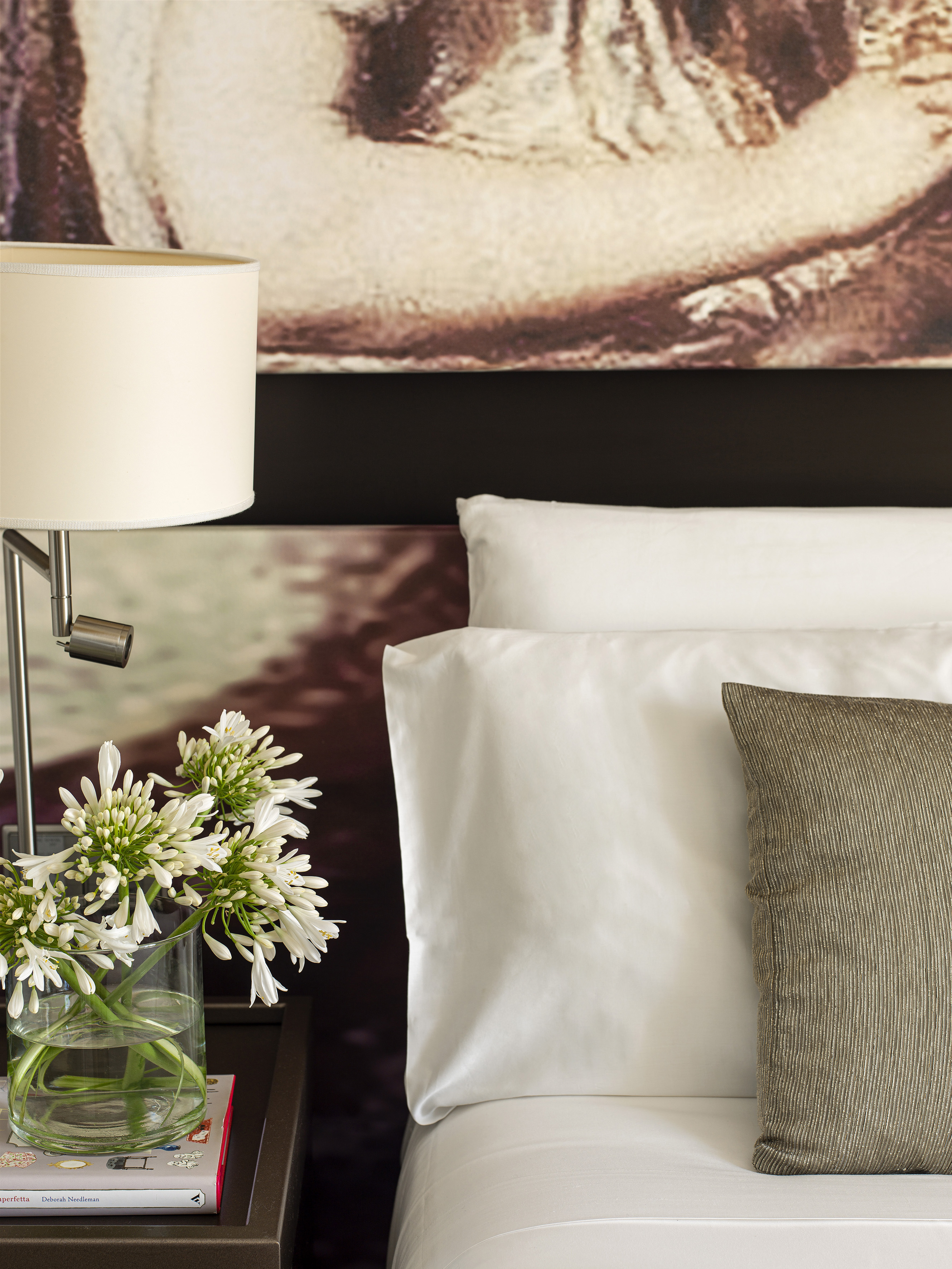 Luxury bedroom with white bedding, striped accent pillow, bedside lamp, and fresh white flowers.