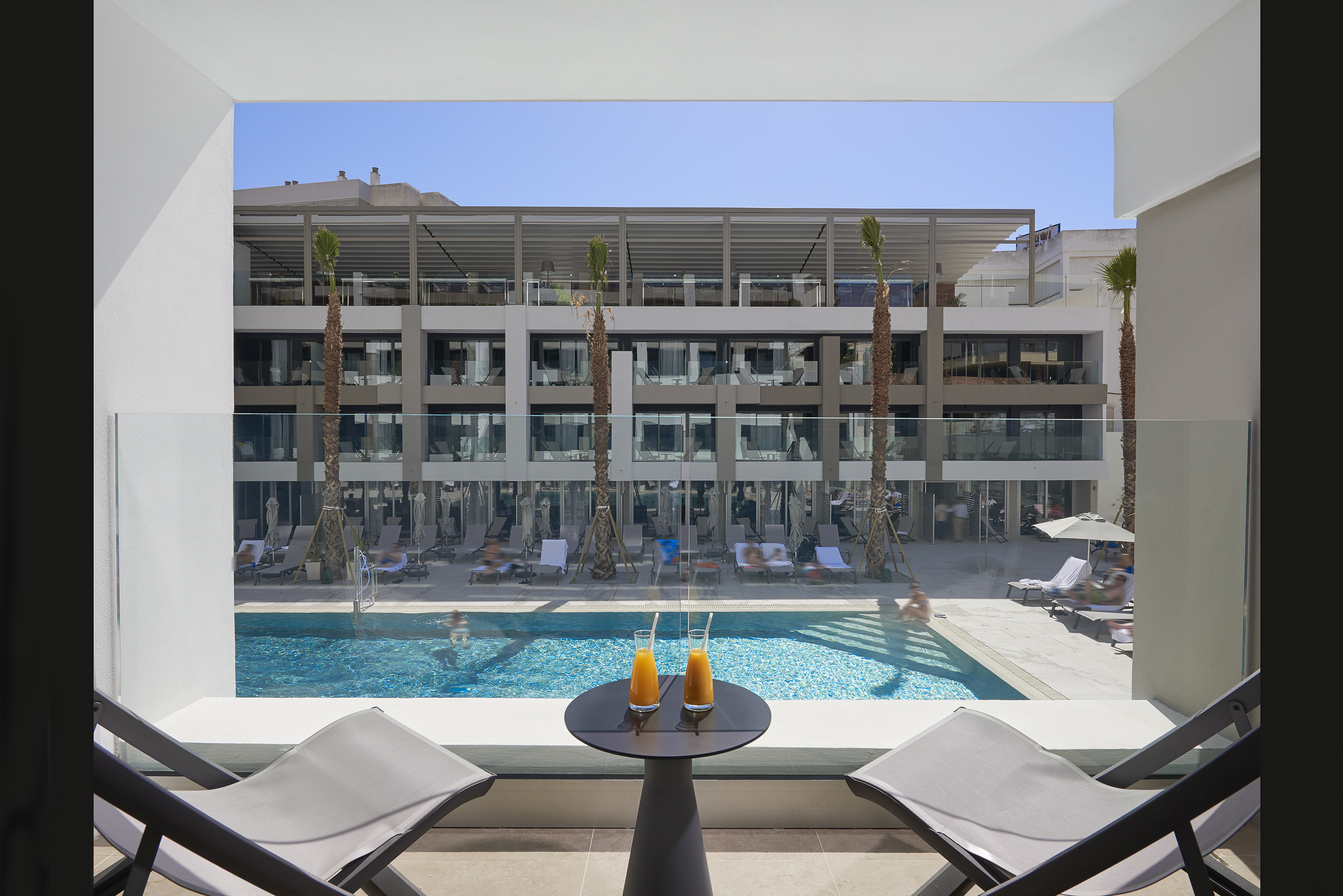 Balcony view of hotel pool, modern building, palm trees, and orange juice.