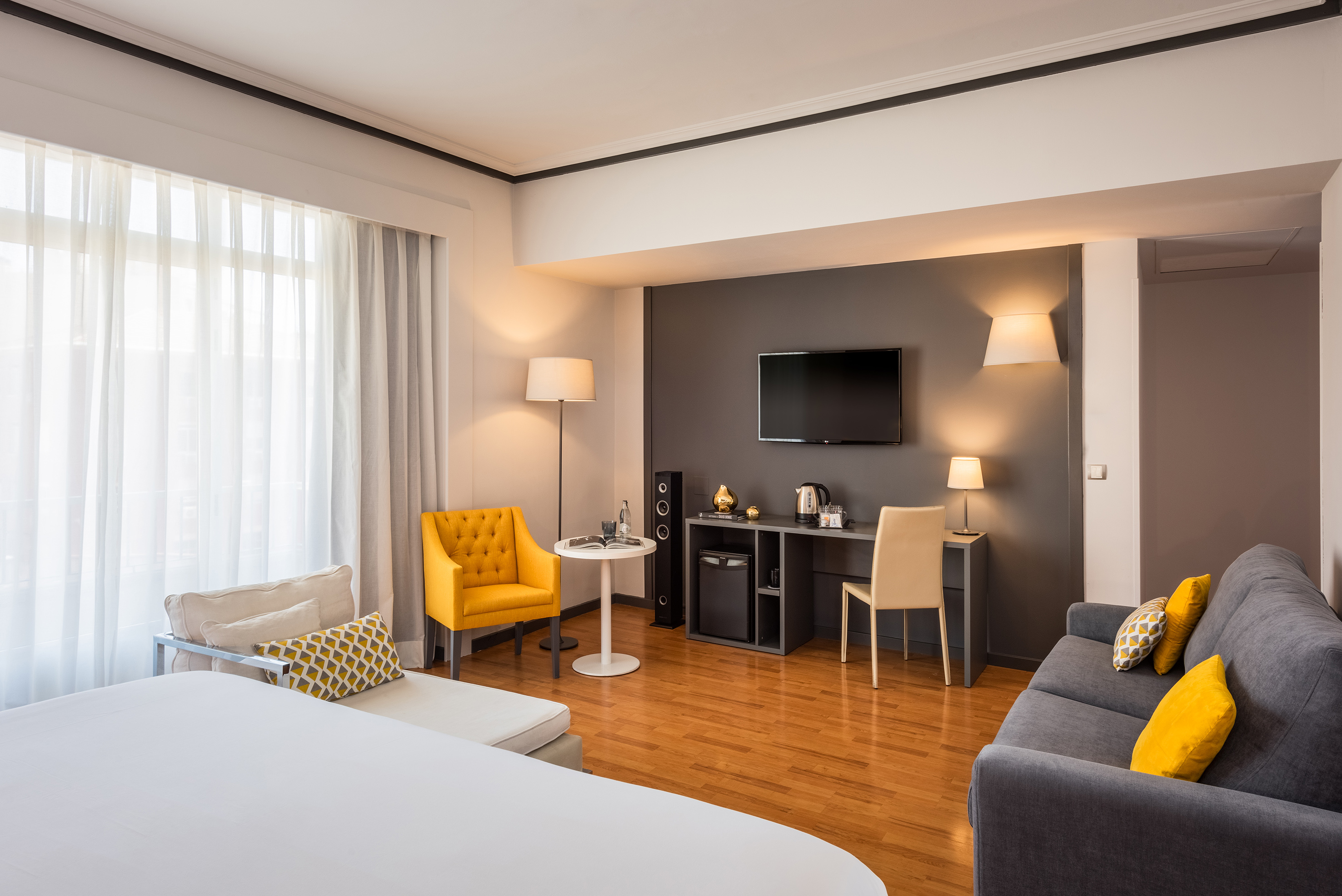 Hotel room interior with bed, grey sofa, yellow armchair, TV, hardwood floor.