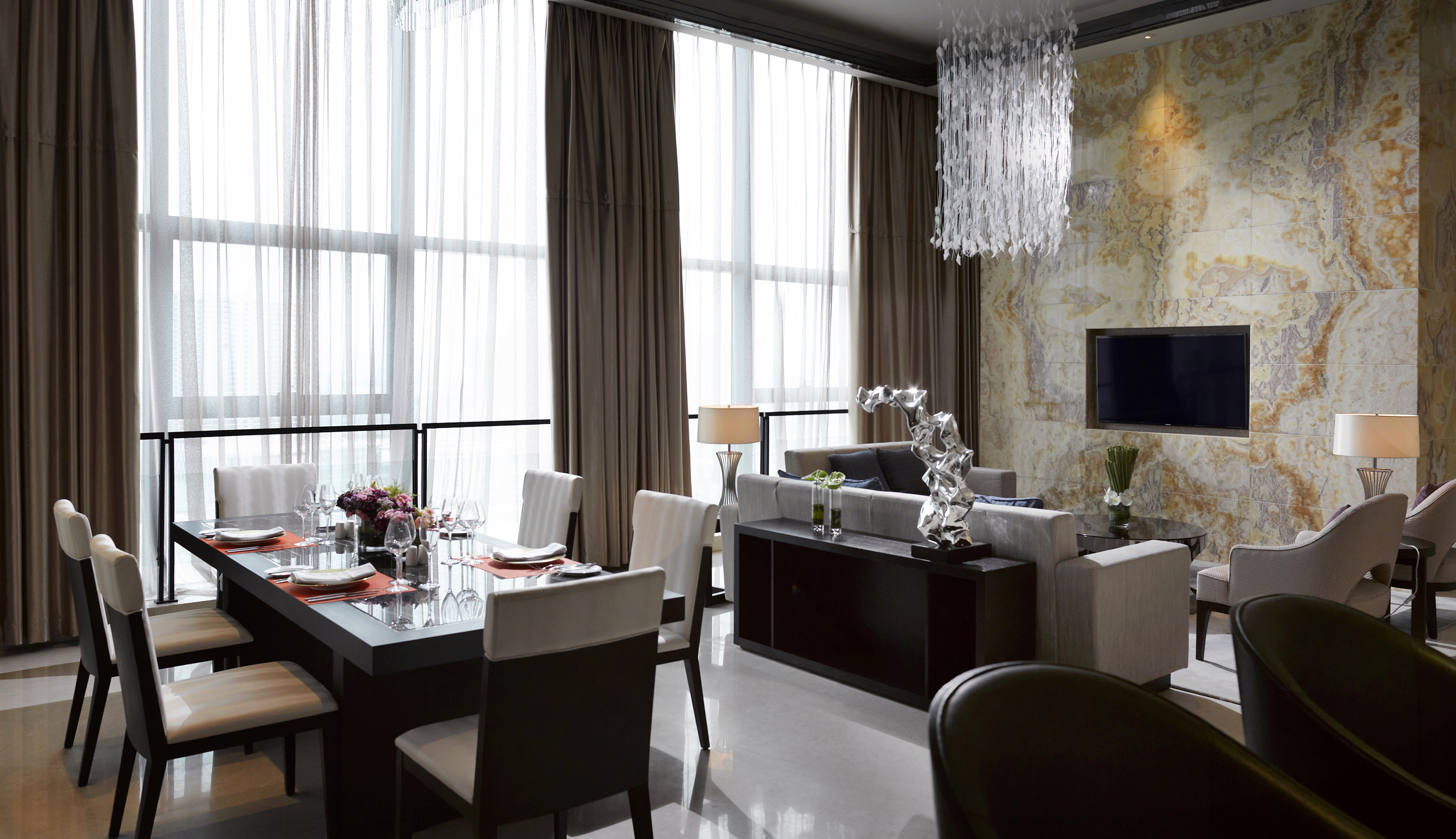 Luxury modern dining and living room with large windows, marble wall, and chandelier.