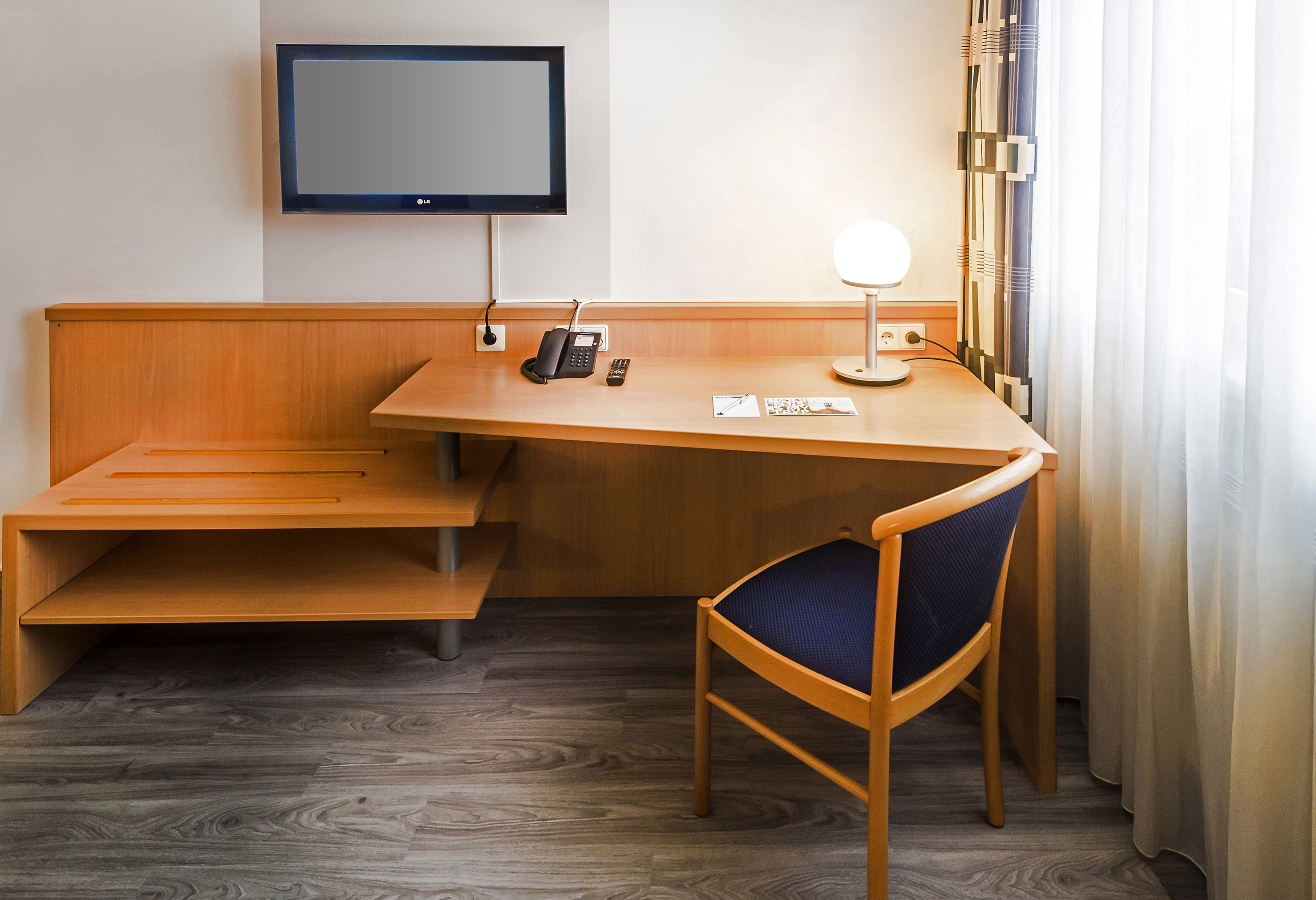 Compact hotel room workstation with wooden desk, TV, lamp, and blue chair.