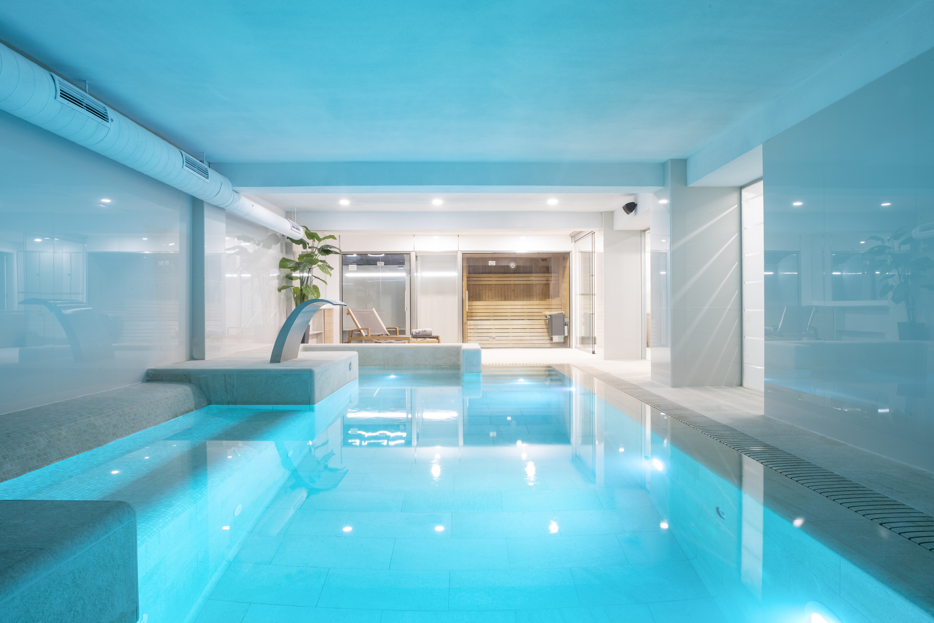 Modern indoor spa pool with blue lighting, waterfall feature, and sauna.