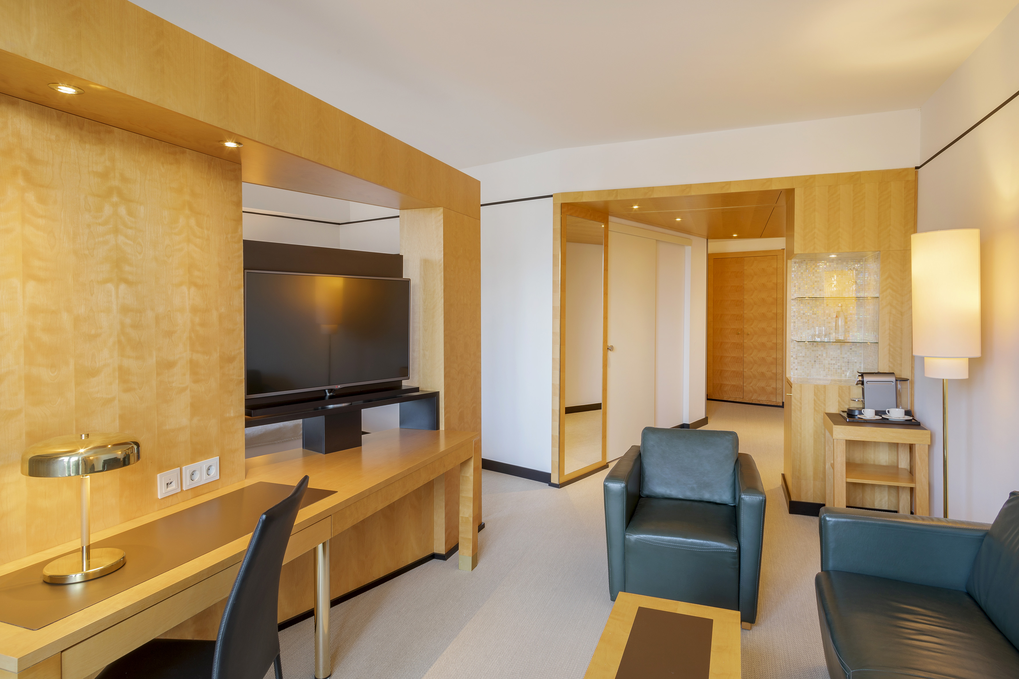 Modern hotel suite with light wood walls, integrated TV, desk, and dark leather armchairs.