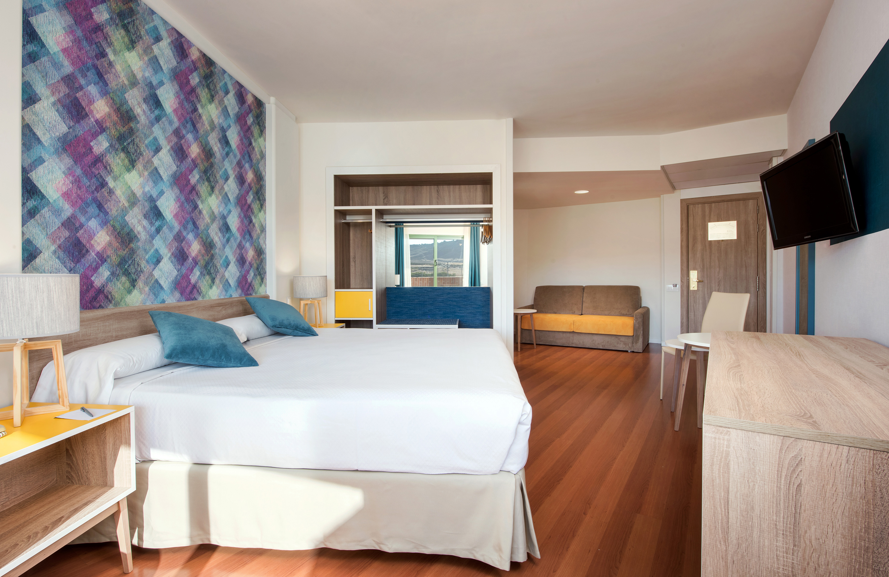 Hotel room with colorful geometric accent wall, bed, sofa, and wood floor.