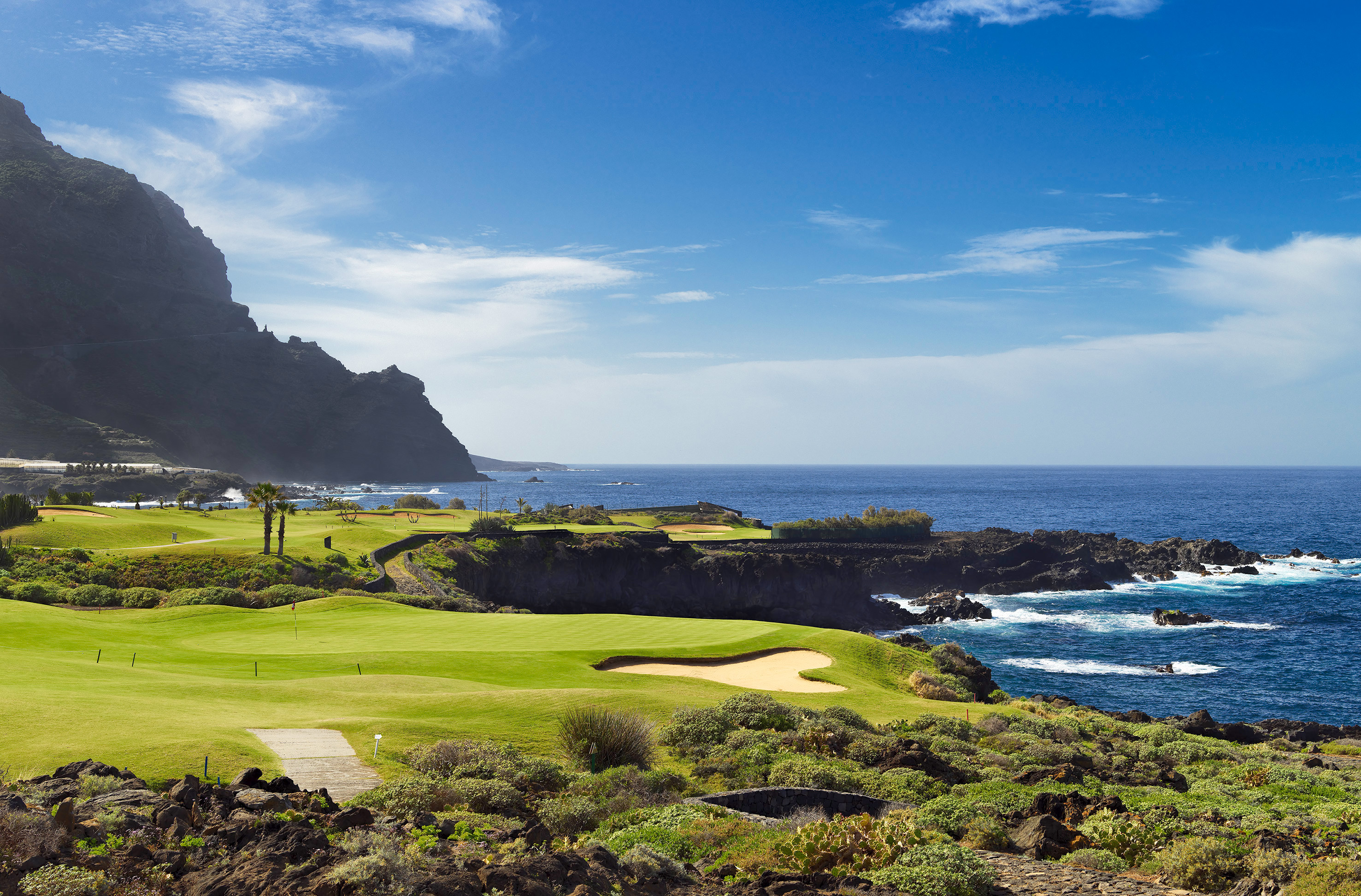 Green golf course by the ocean, rocky coast, and large cliffs under blue sky.