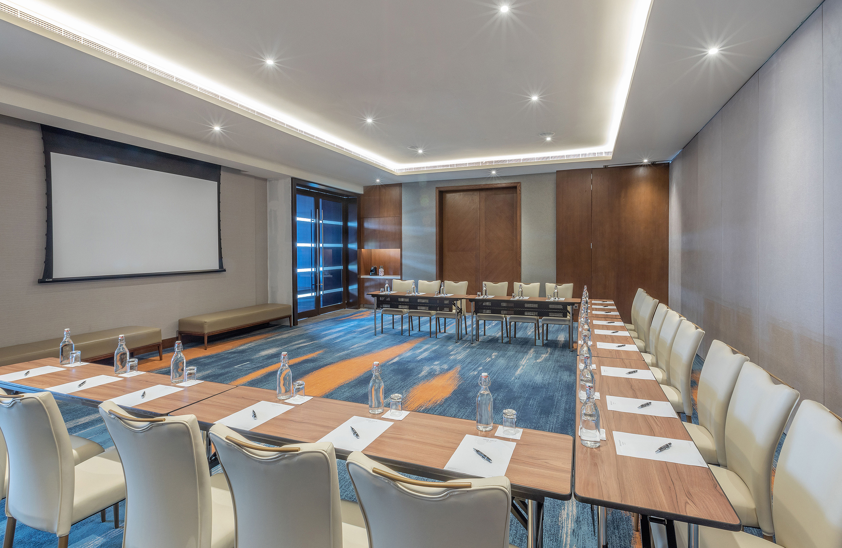 Modern meeting room with U-shaped setup, projector screen, patterned carpet.