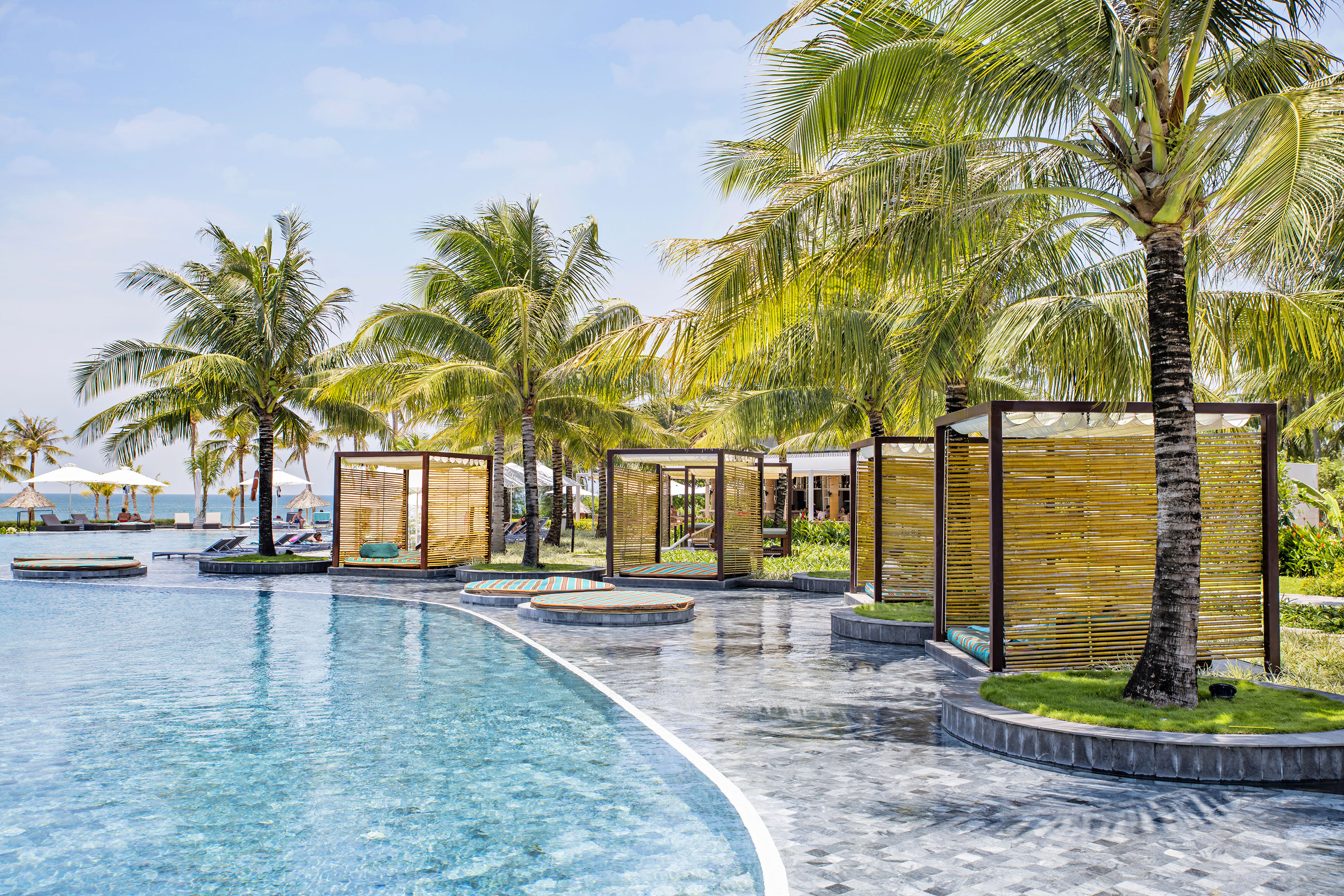 Tropical resort swimming pool with bamboo cabanas and lush palm trees.