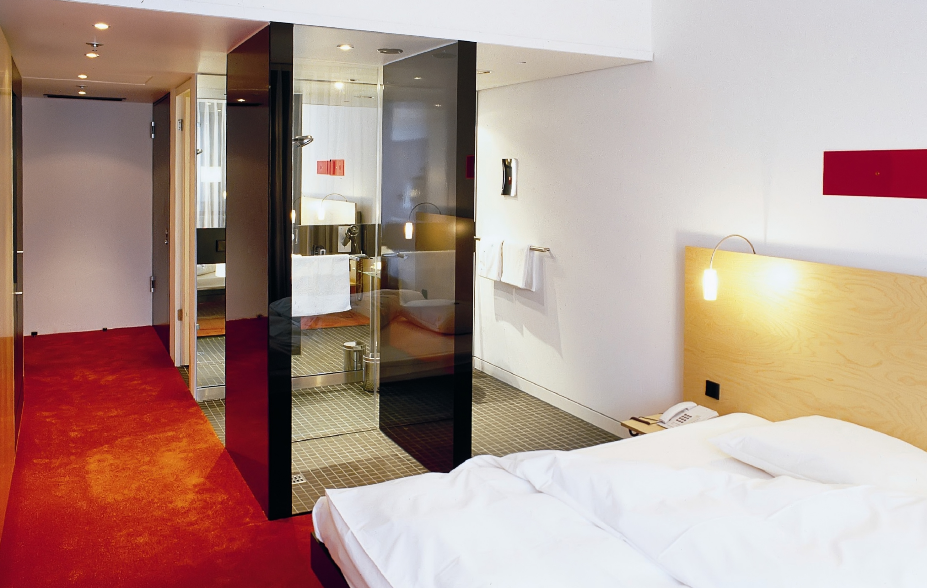 Modern hotel room with red carpet, glass-walled bathroom, and white bed.