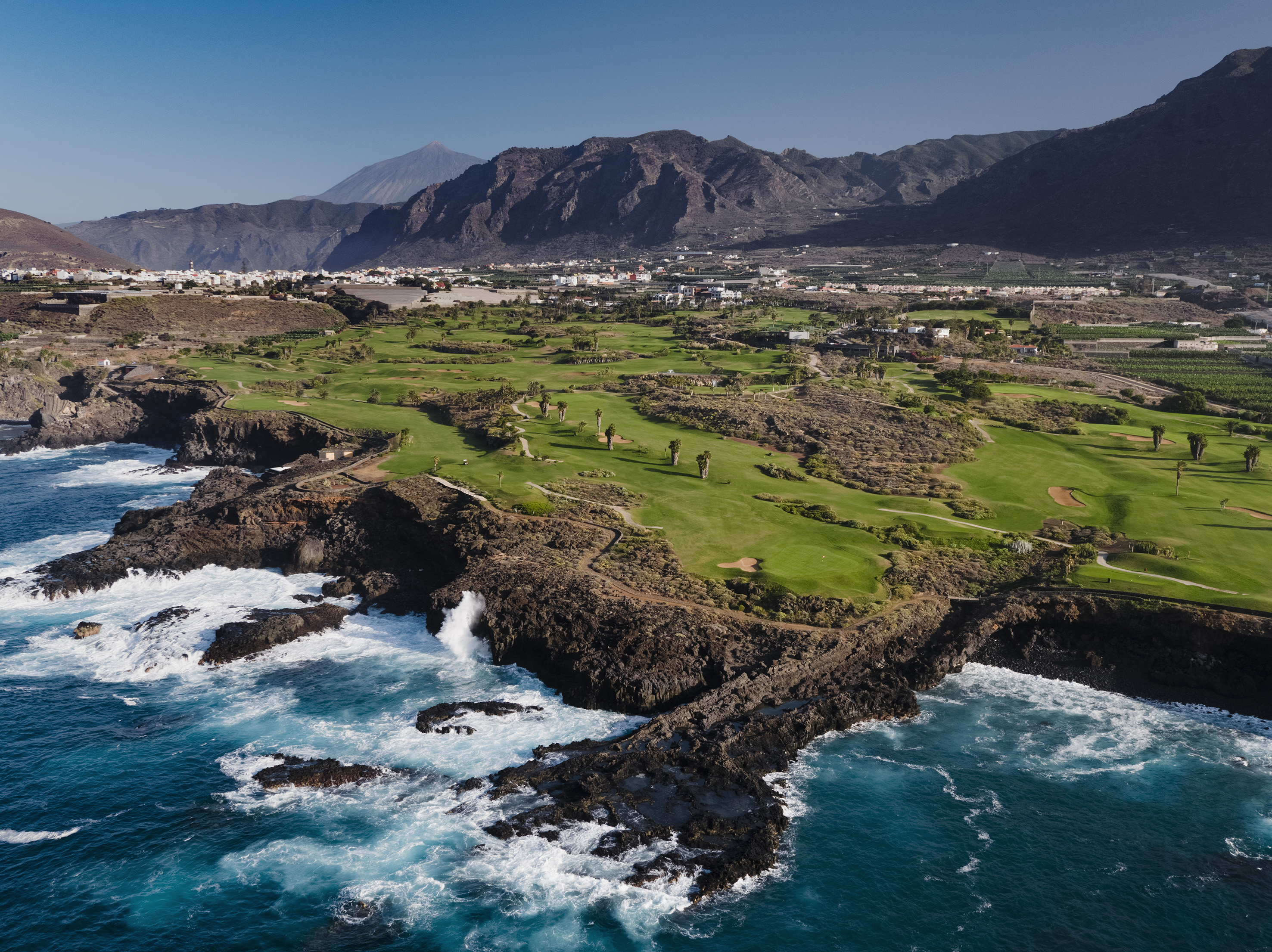 Aerial coastal golf course, Tenerife, with ocean, volcanic mountains, and town.