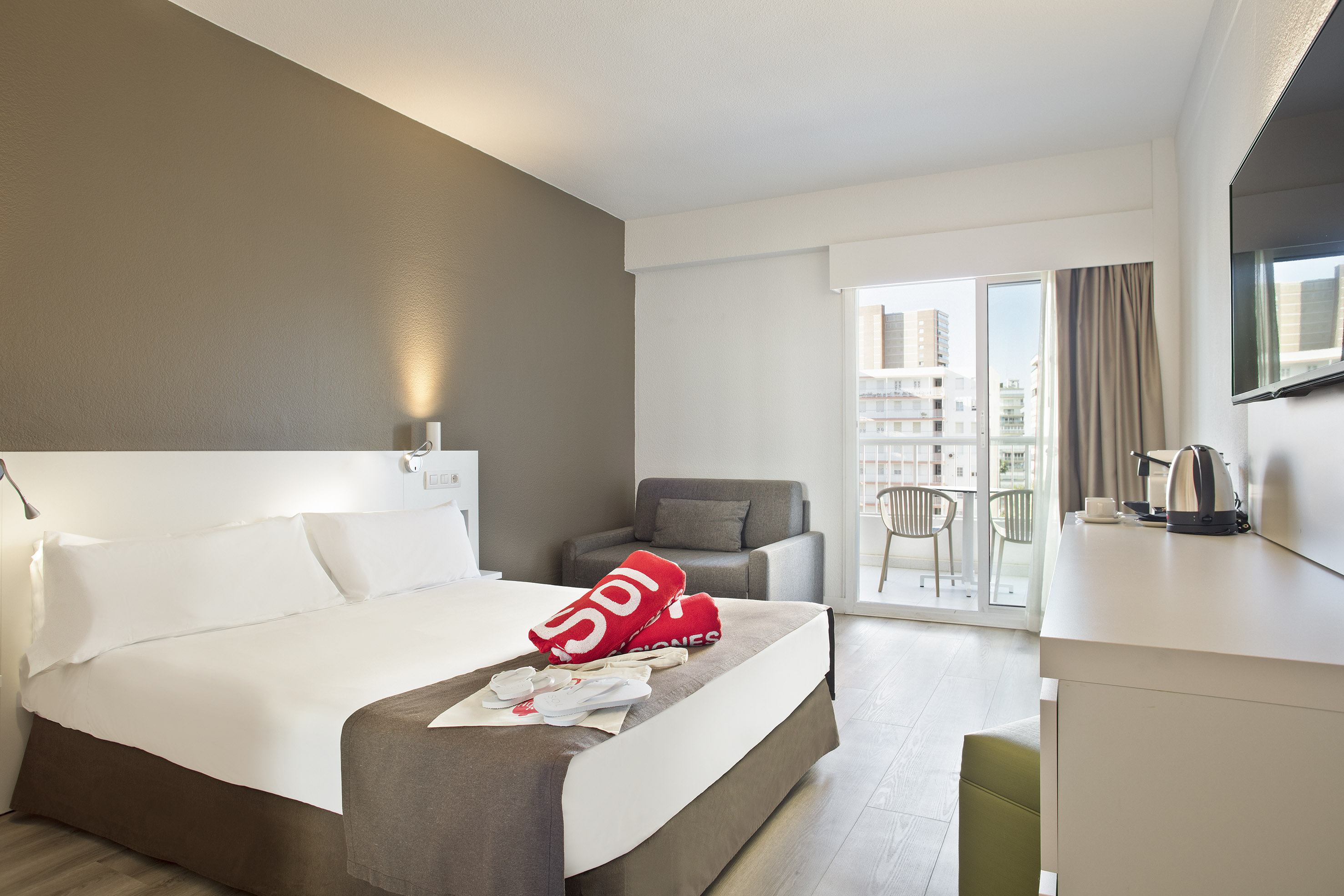 Modern hotel room with bed, sofa, balcony city view. Red towels & slippers on bed.