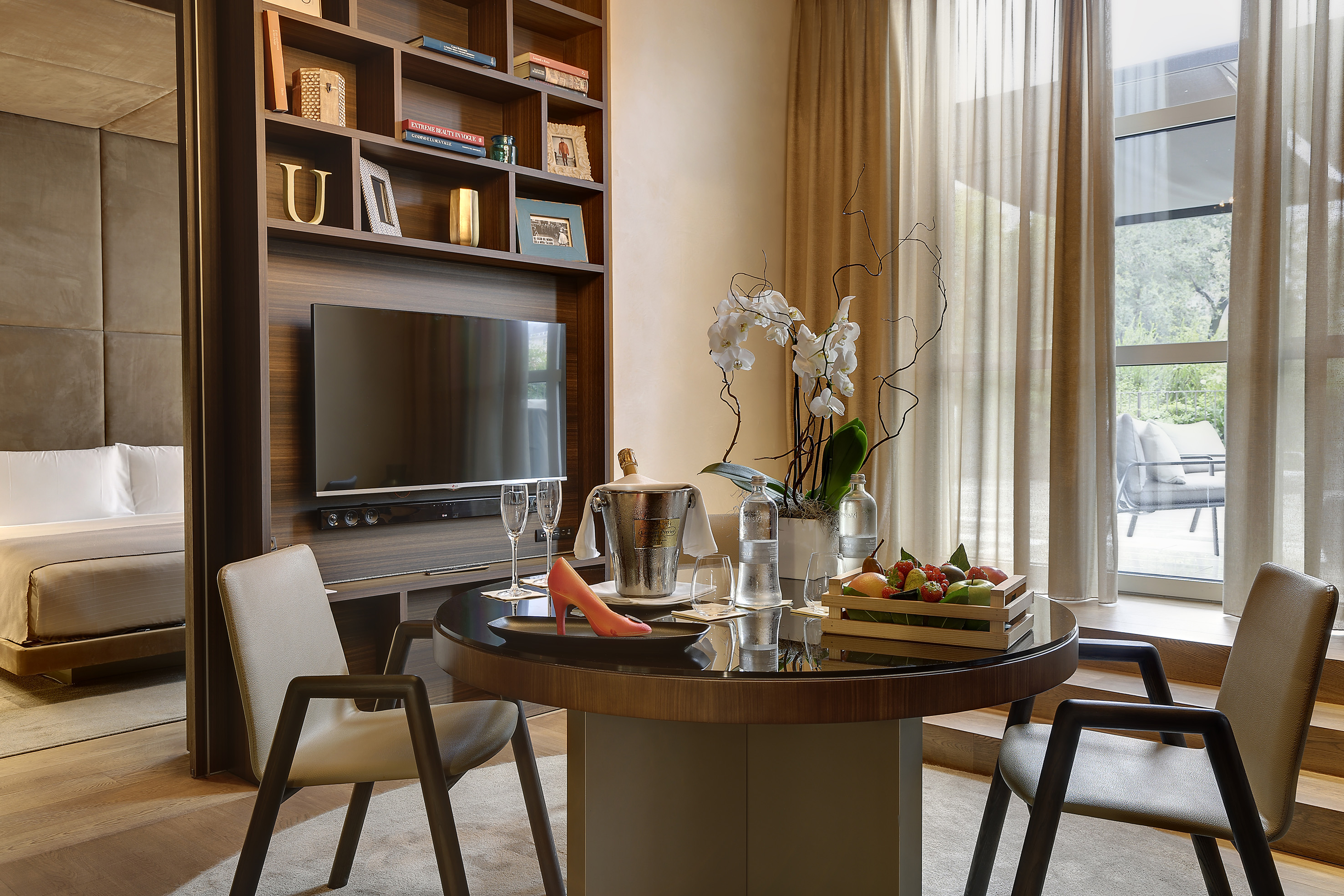 Modern hotel suite: dining table with champagne, fruit, orchids, and bed.