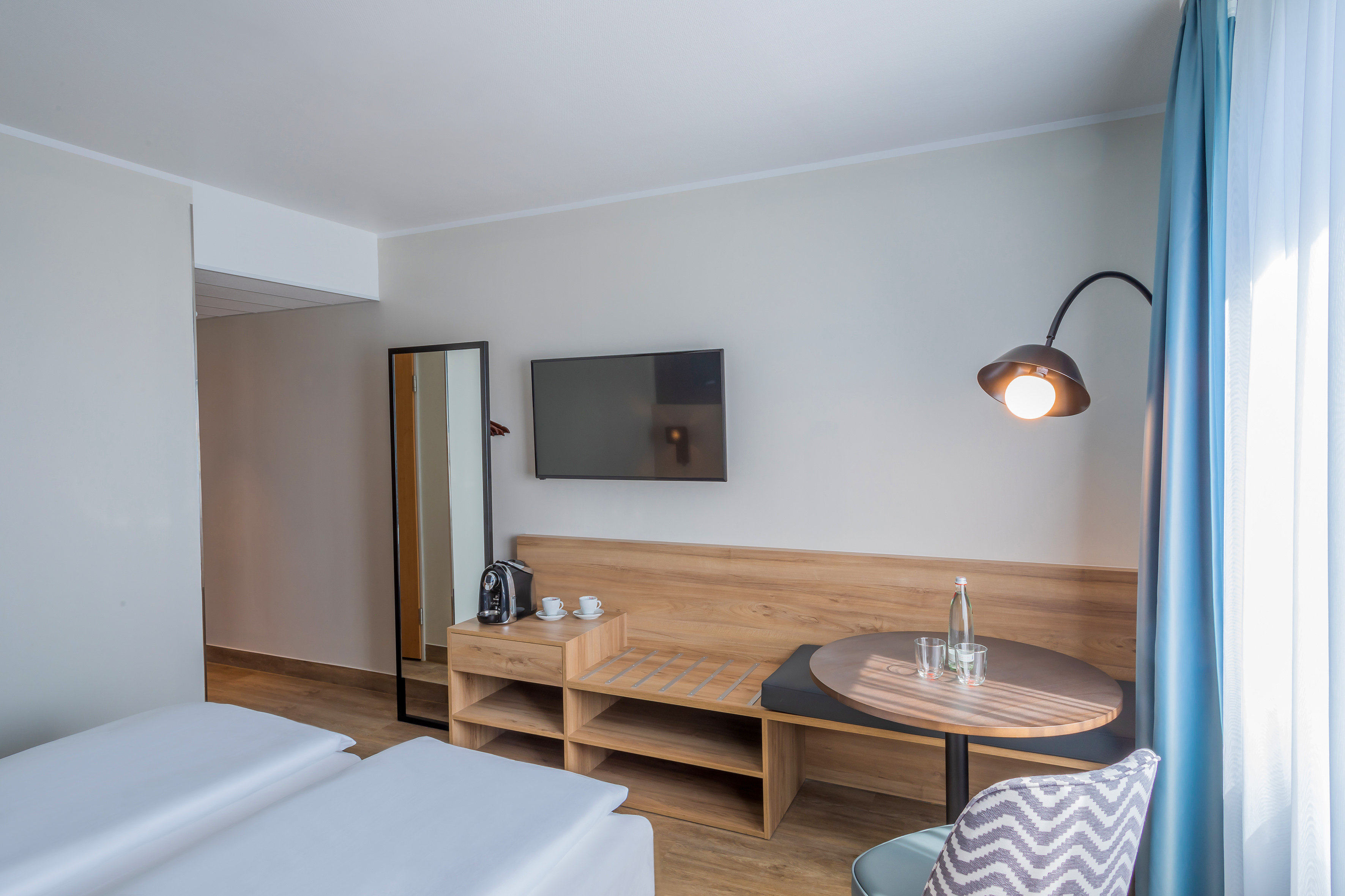 Modern hotel room with twin beds, wall TV, coffee maker, and dining table.